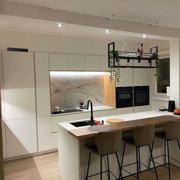 Modern kitchen with white cabinets, a marble backsplash, and a kitchen island with three chairs. There is a hanging shelf with bottles and greenery above the island, and built-in ovens in the cabinetry.