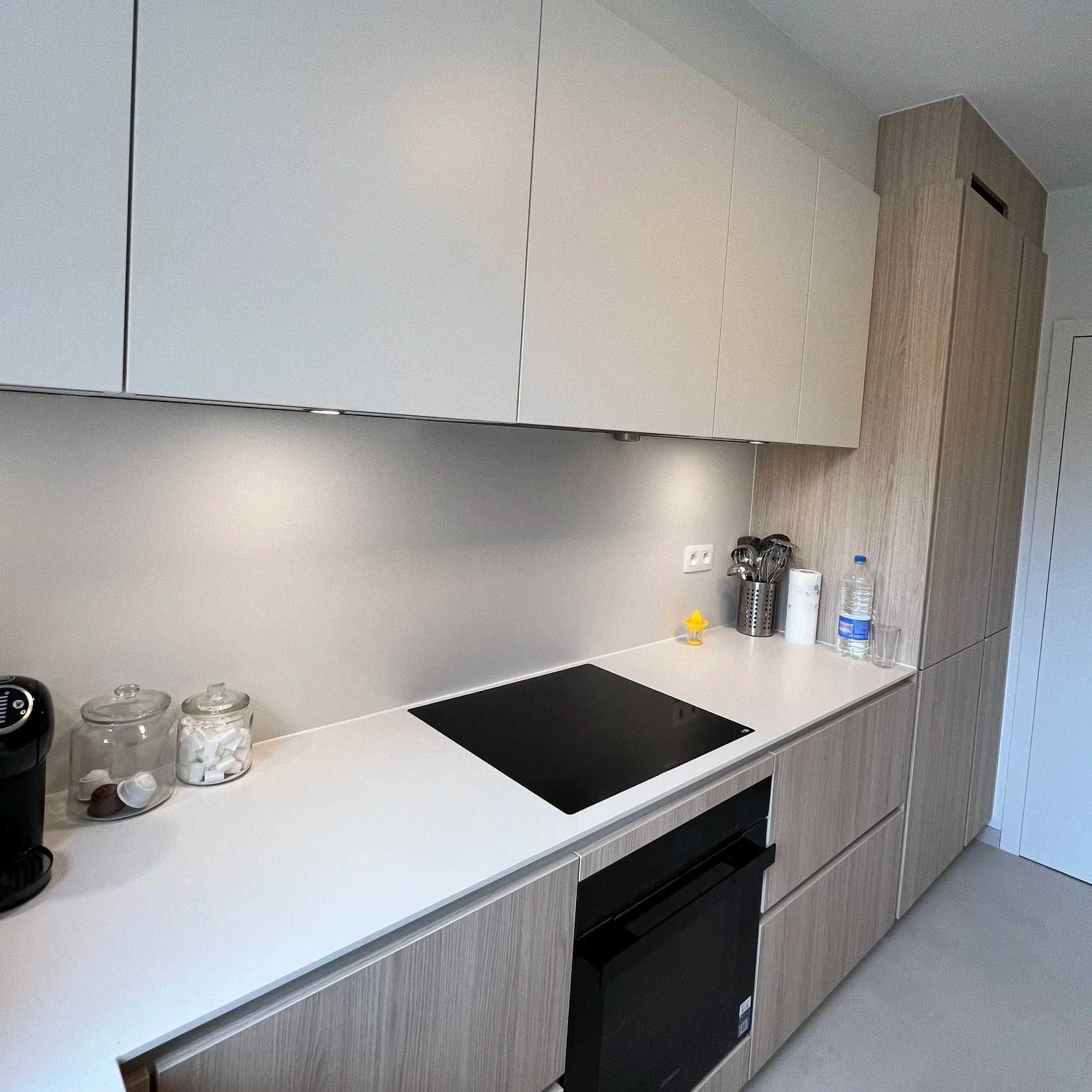A modern kitchen with white and light wood cabinets, a white countertop, a black stovetop, and various kitchen items such as jars, a coffee machine, utensils, a bottle of water, and paper towels.