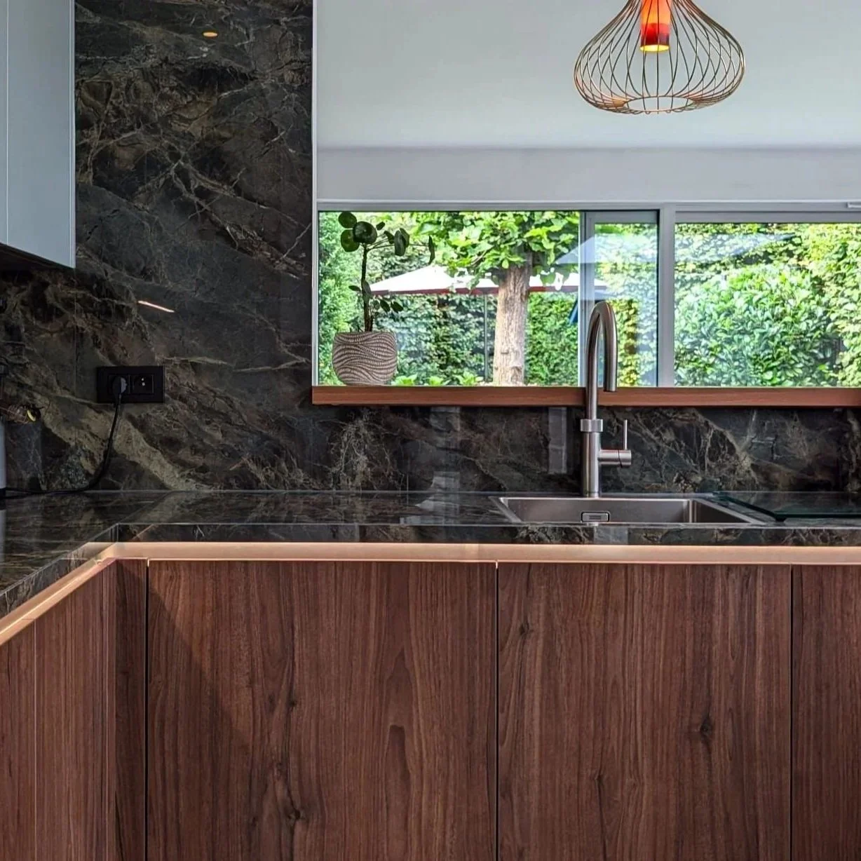 Modern kitchen with dark marble backsplash and countertop, wooden lower cabinets, a stainless steel sink with a high-arc faucet, a large window overlooking greenery outside, a potted plant on the windowsill, and a decorative wooden pendant light hanging from the ceiling.