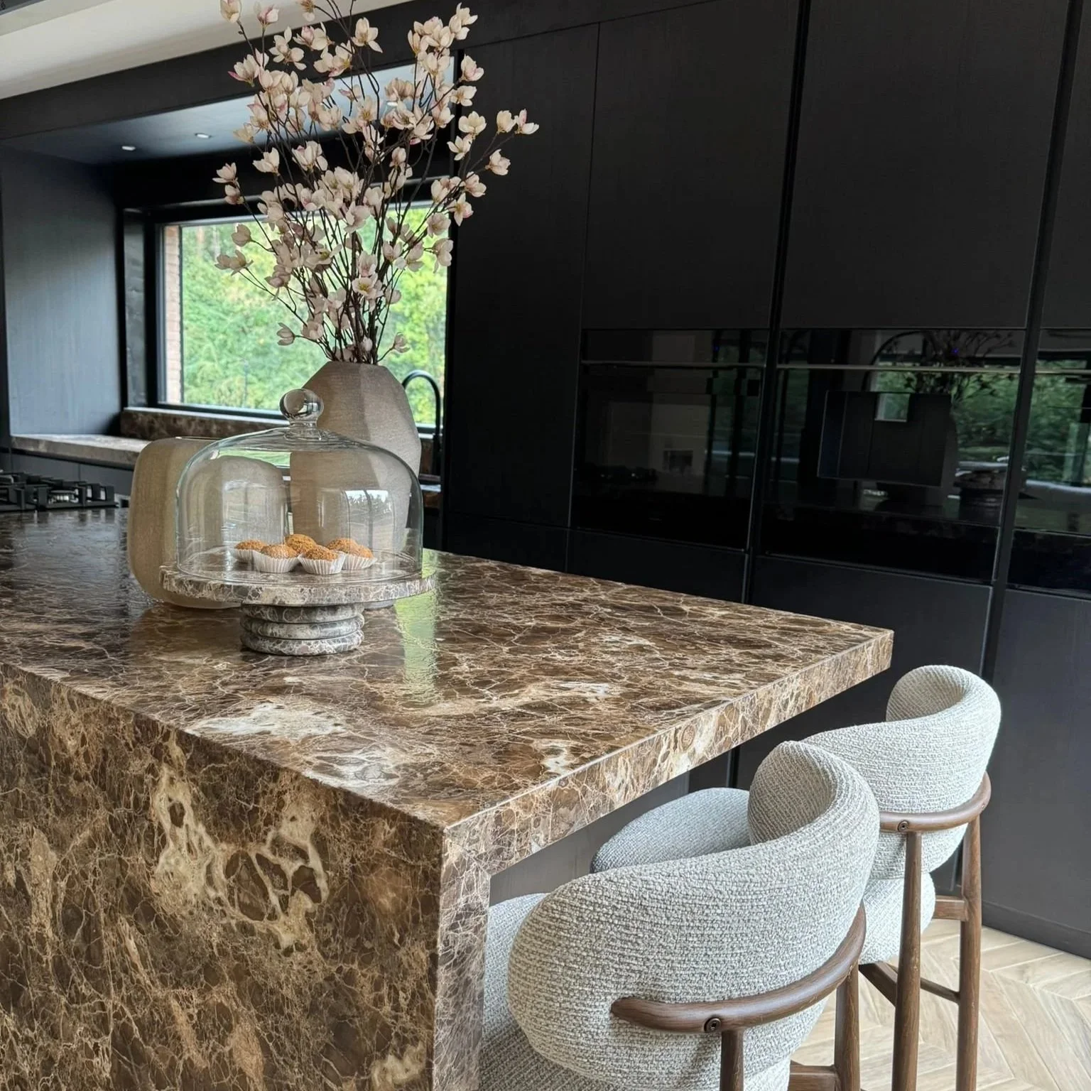 Modern kitchen with a marble island, a vase of pink flowers, and a glass cake dome with pastries, with black cabinetry and a window overlooking greenery.