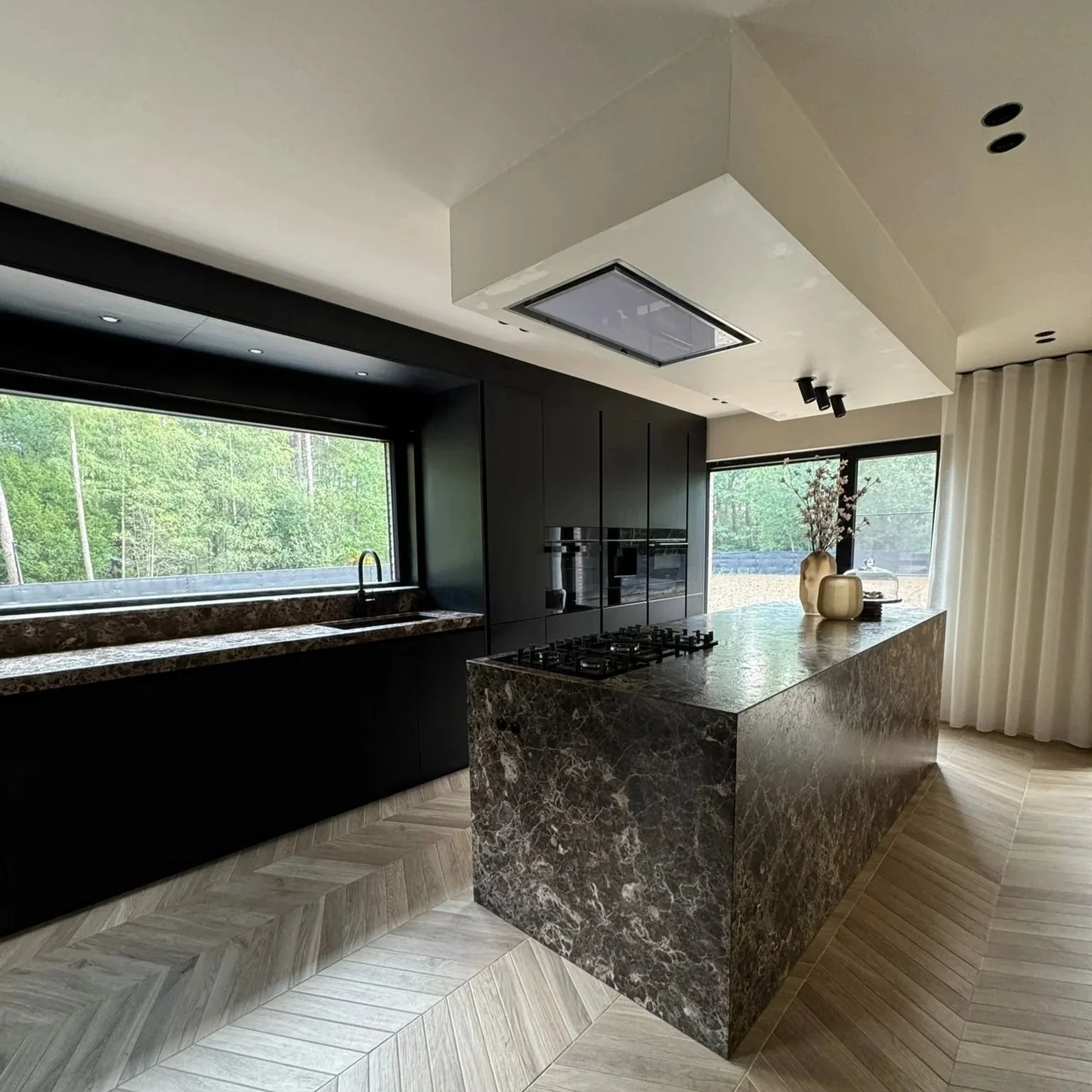 Modern kitchen with black cabinetry, a marble island, and large windows showing a green outdoor landscape.