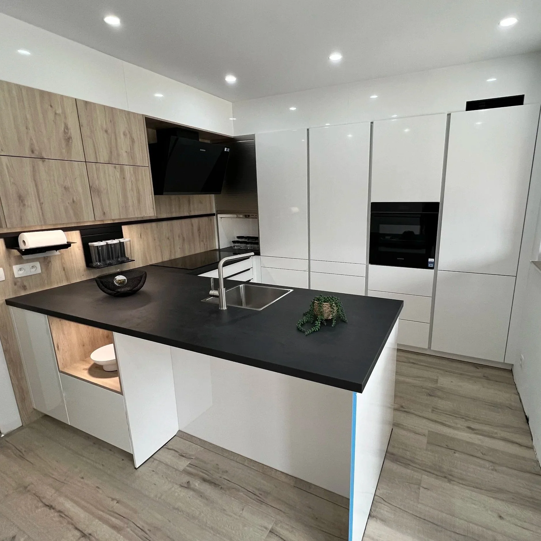 Modern kitchen with white cabinets, black countertop island with sink, wooden accent cabinets, built-in appliances, and wooden flooring.