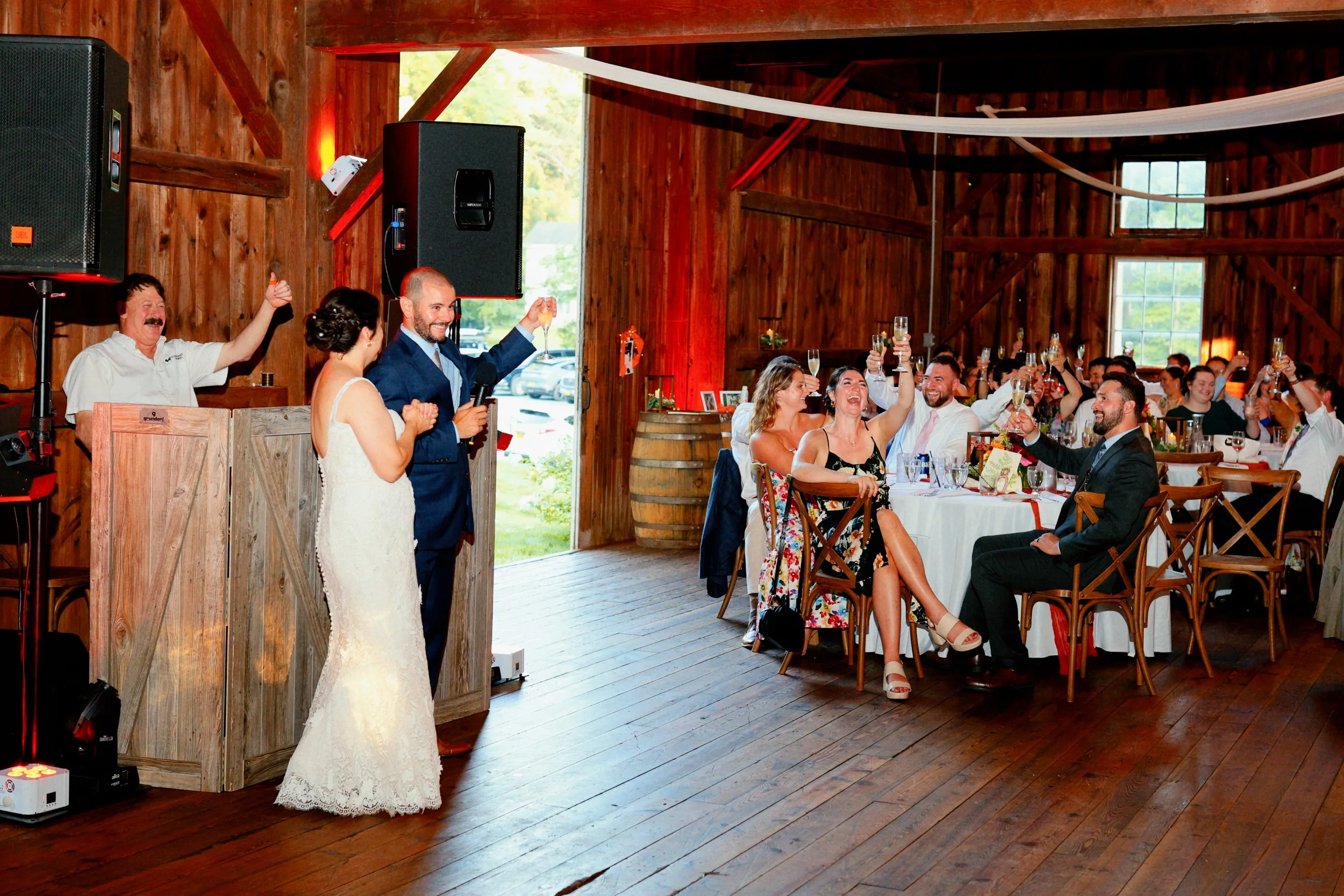 A wedding reception inside a rustic wooden barn with guests raising glasses in a toast while the bride and groom watch and smile.