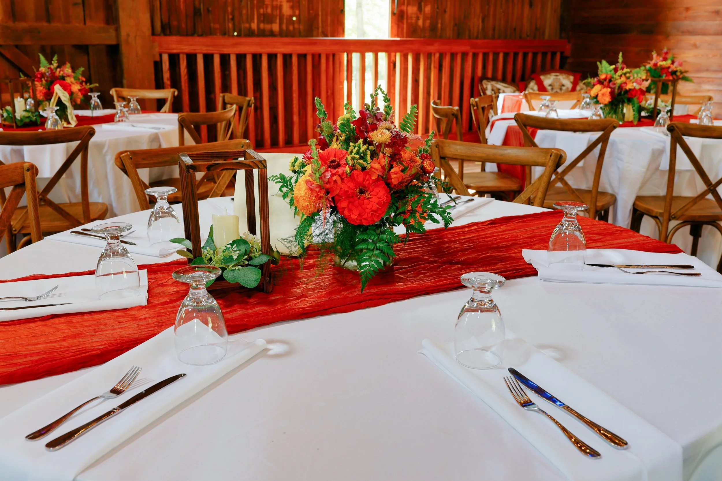 Elegant dining table setup with white tablecloths, red runners, floral centerpieces, and glassware in a rustic wooden interior.