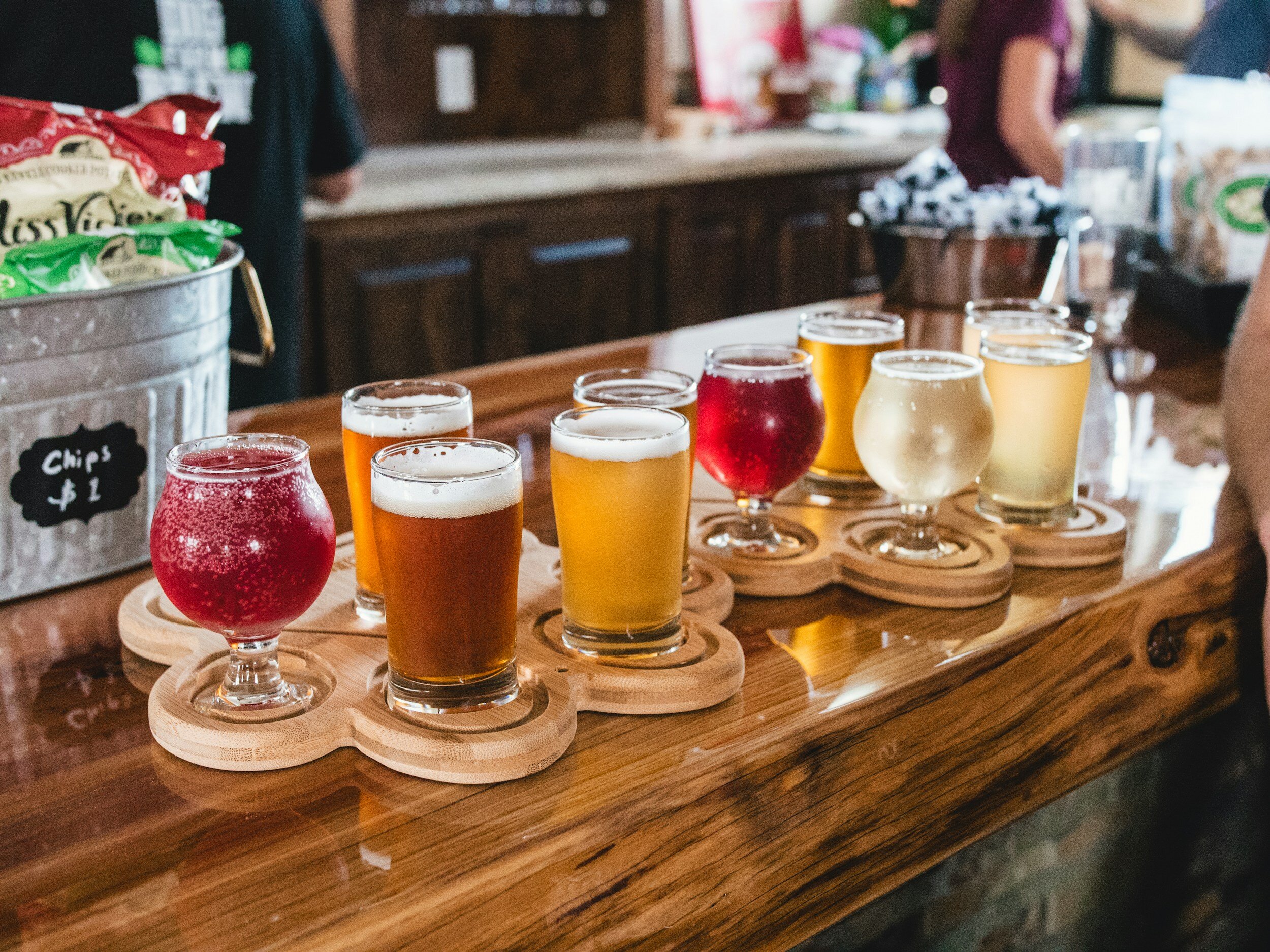 A flight of beers on a wooden tray with various types and colors, placed on a bar counter next to a bucket of chips and other items.
