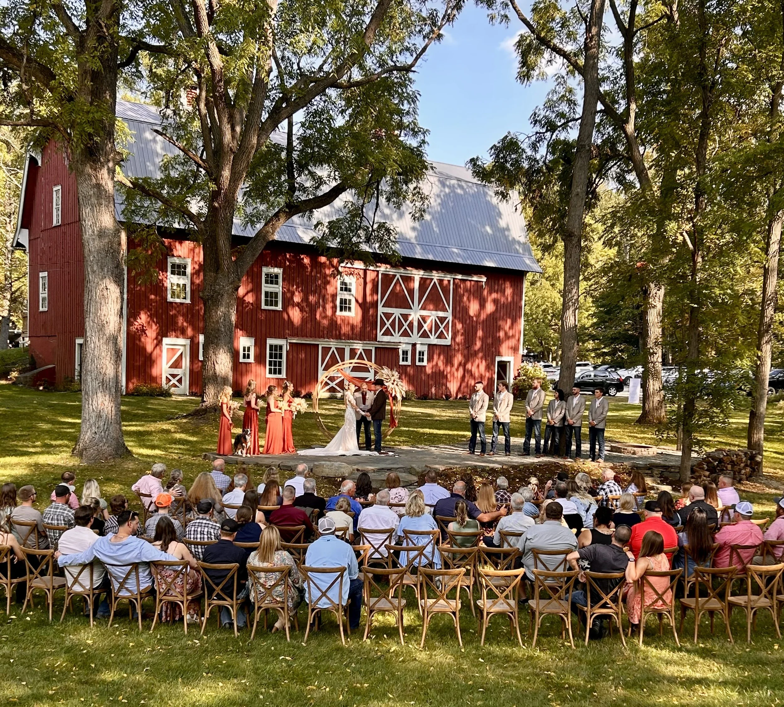 People attending an outdoor wedding ceremony with a red barn in the background and surrounded by large trees.