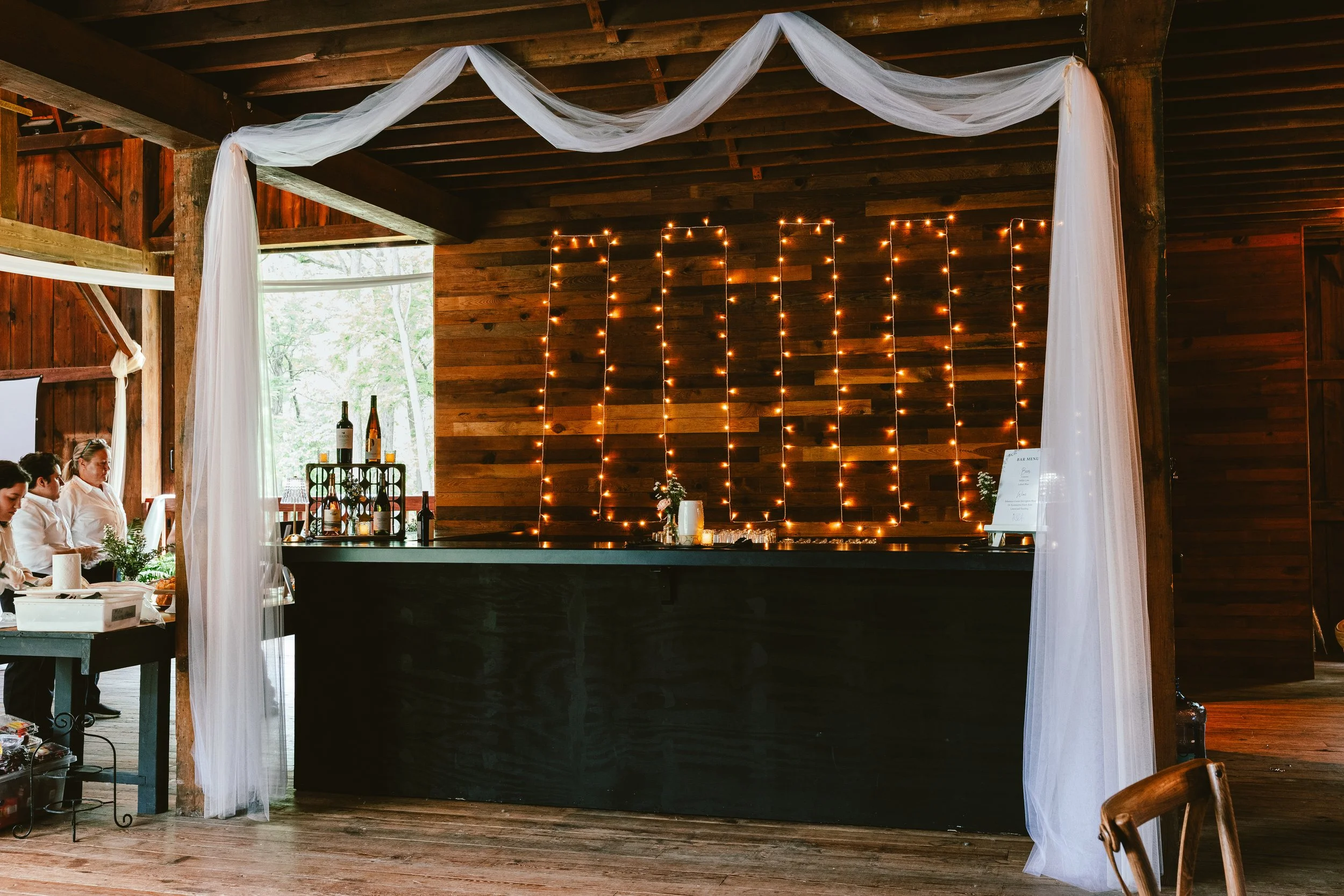 Interior of a rustic wooden venue with string lights and white drapery decor, likely for a celebration or wedding, with a bar area and guests seated on the left.