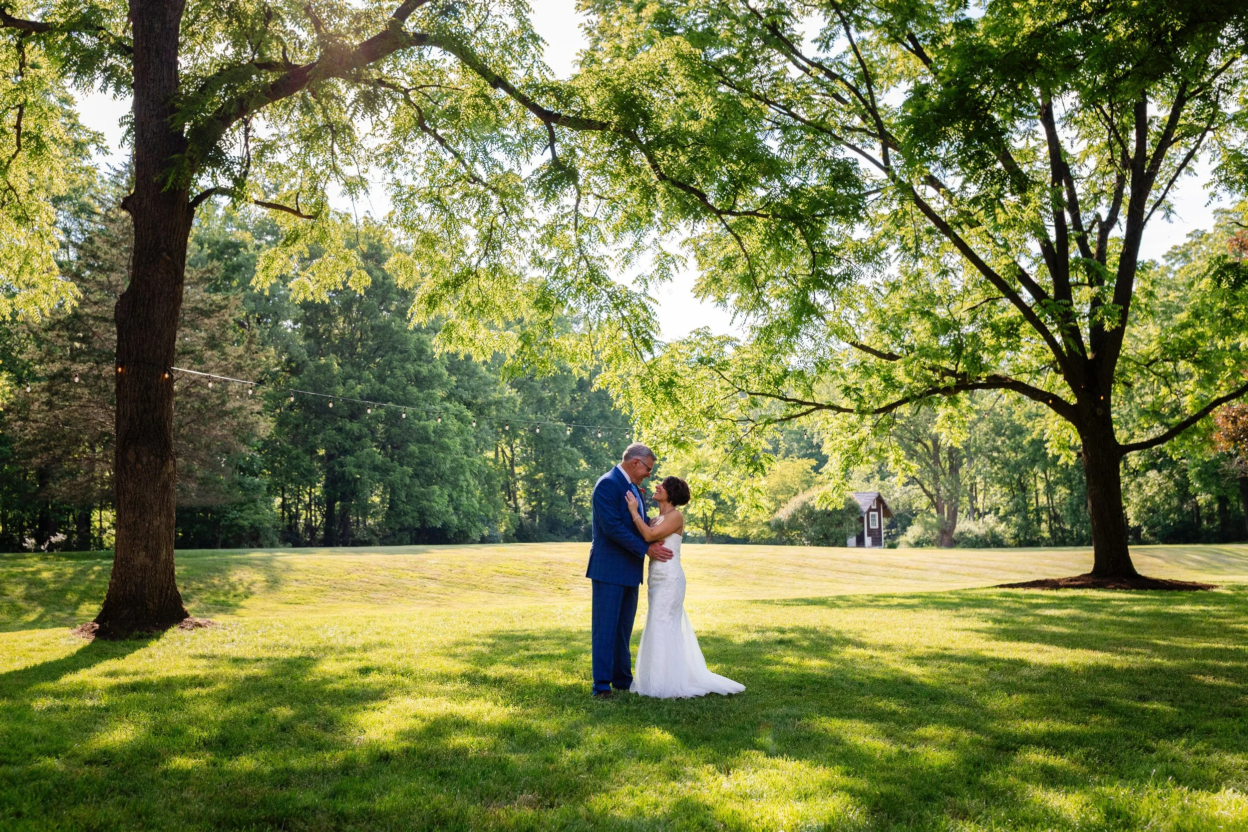 A bride and groom standing together in a grassy park, surrounded by large trees, with sunlight filtering through the leaves, during their wedding.