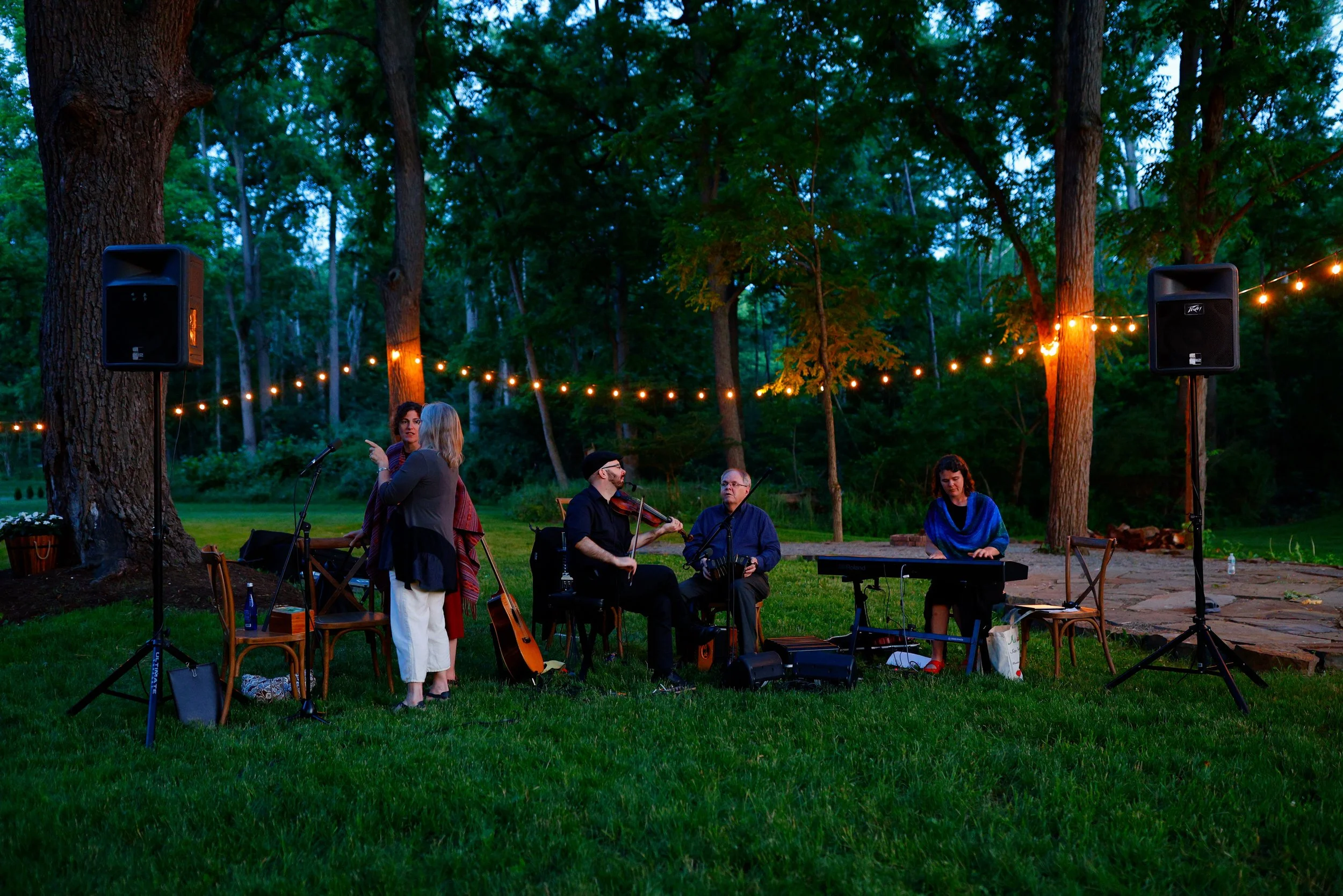 A group of musicians performing outdoors under string lights in a wooded area during dusk.
