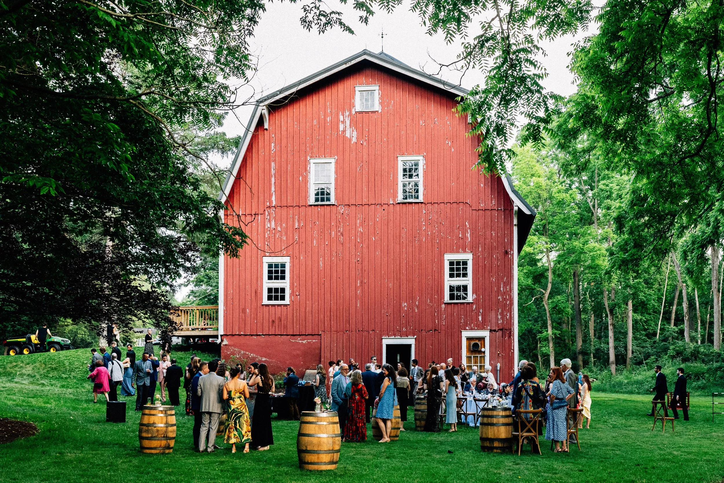 People gathered outside a large red barn for a social event, with tables, chairs, and barrels on a grassy lawn surrounded by trees.