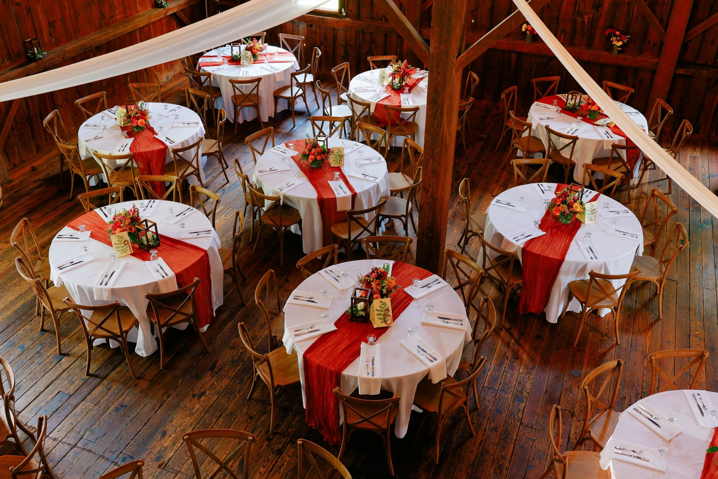 Wedding reception setup inside a rustic wooden venue with round tables decorated with white tablecloths and red runners, floral centerpieces, and wooden chairs.