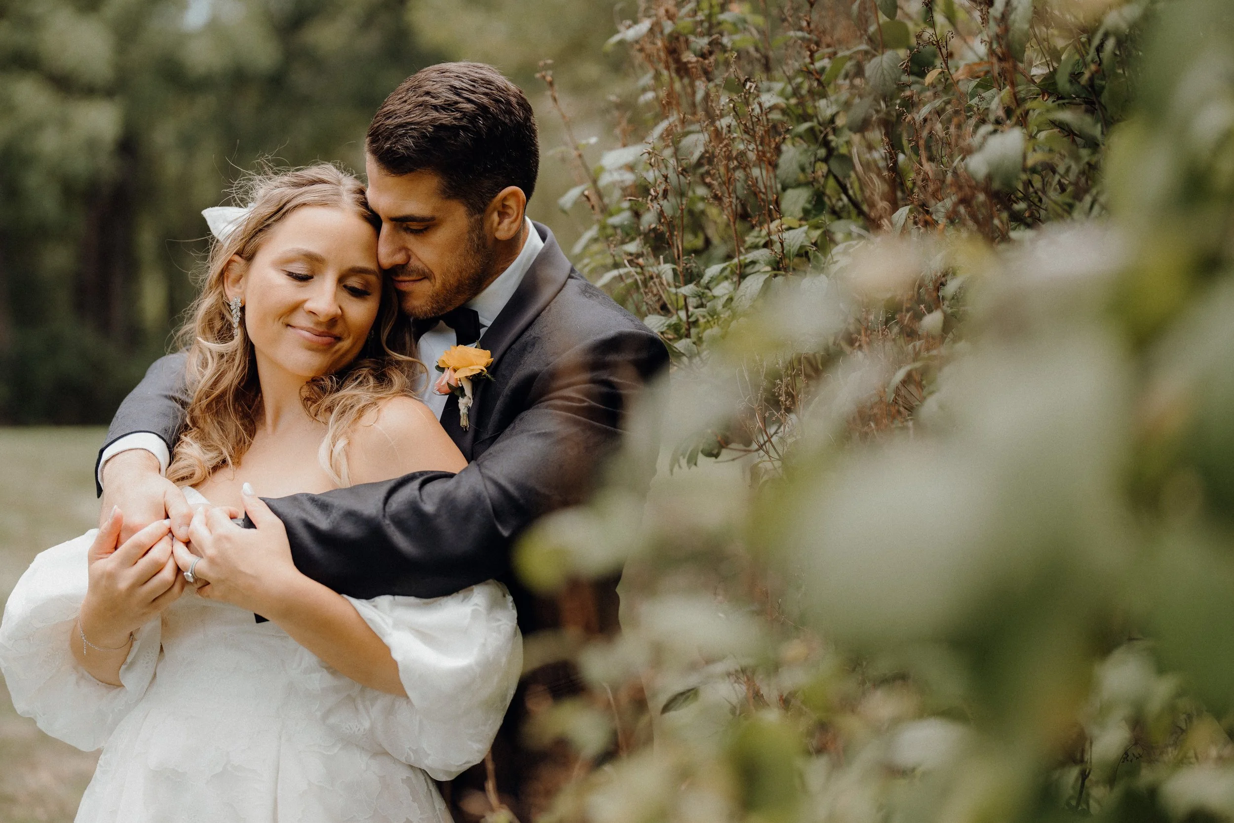 A bride and groom hugging outdoors, both smiling softly with eyes closed, surrounded by greenery.