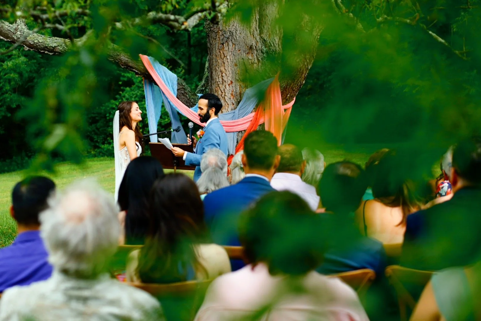 A couple exchanging vows during an outdoor wedding ceremony beneath a large tree adorned with colorful fabric. Guests are seated and watching in the foreground.