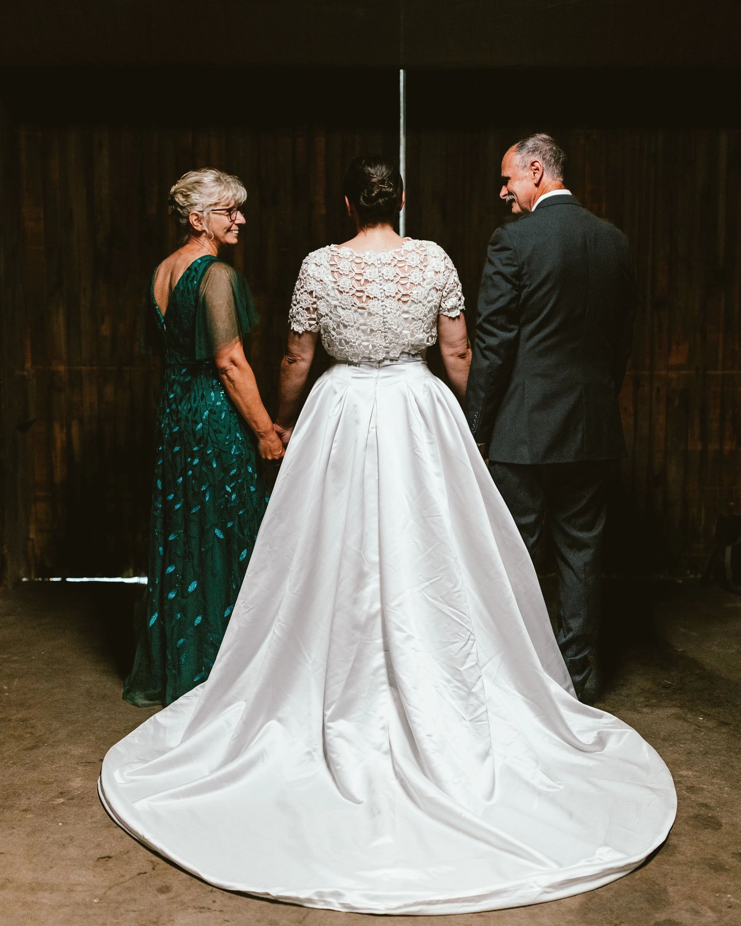 A bride in a white wedding dress facing her parents in front of a dark background.