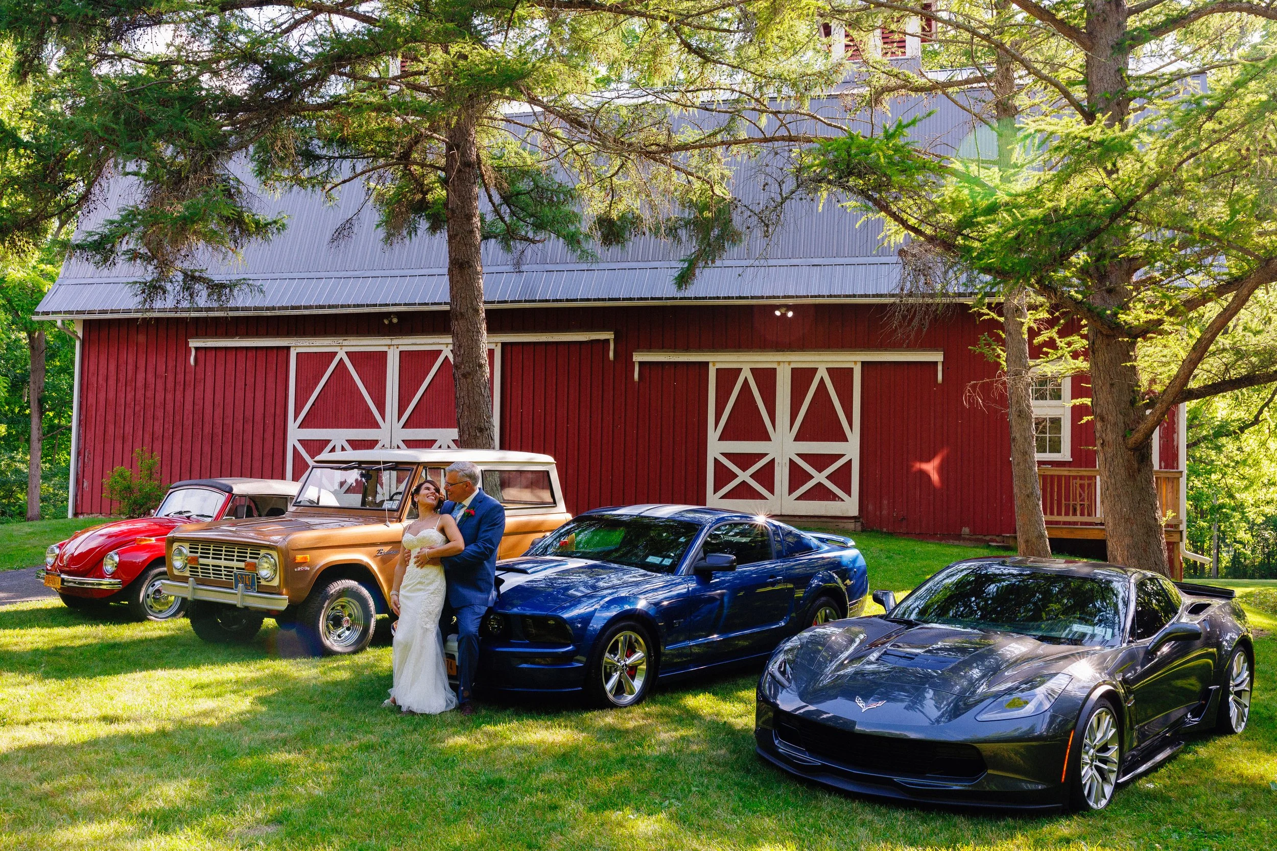 A bride and groom dancing in front of a line of vintage and modern cars on a grassy area outside a red barn surrounded by green trees.
