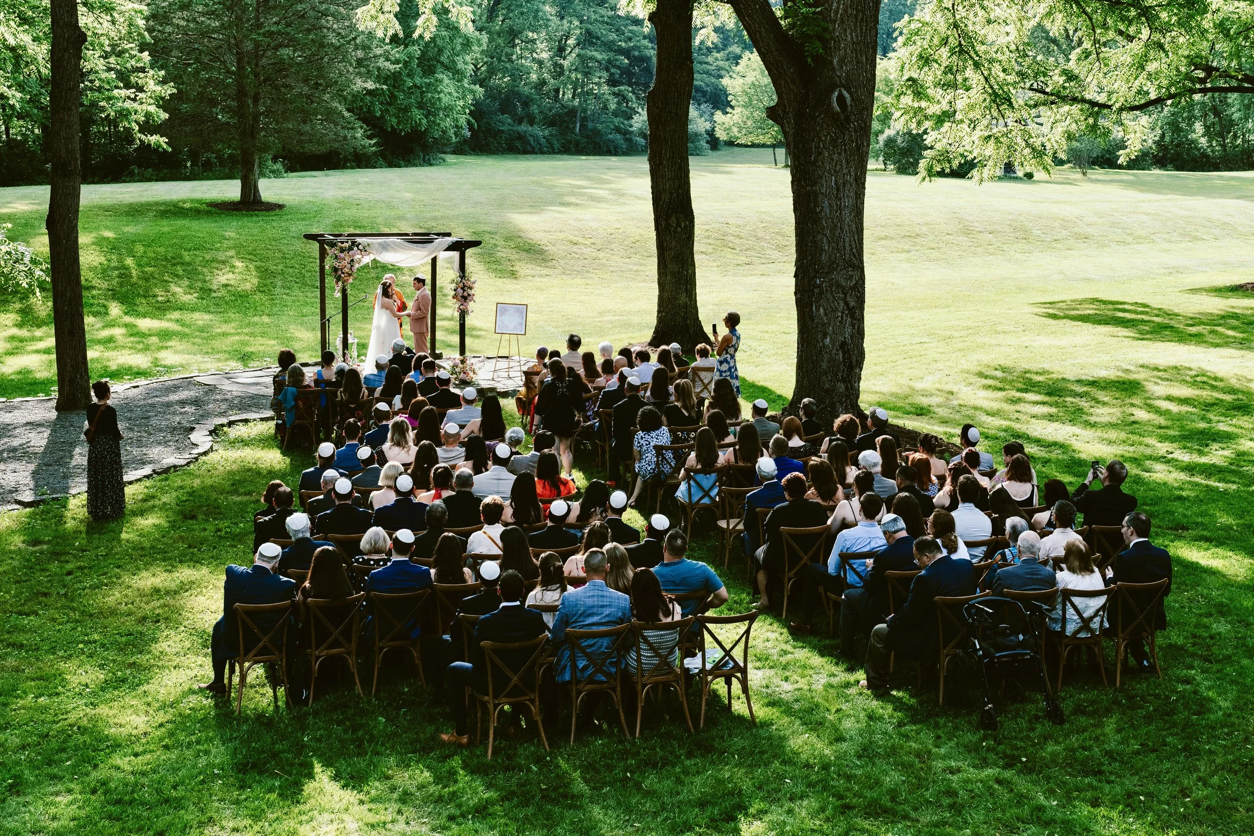 A large group of people gathered outdoors for a wedding ceremony on a grassy lawn under trees, with the bride and groom standing at an altar decorated with flowers.