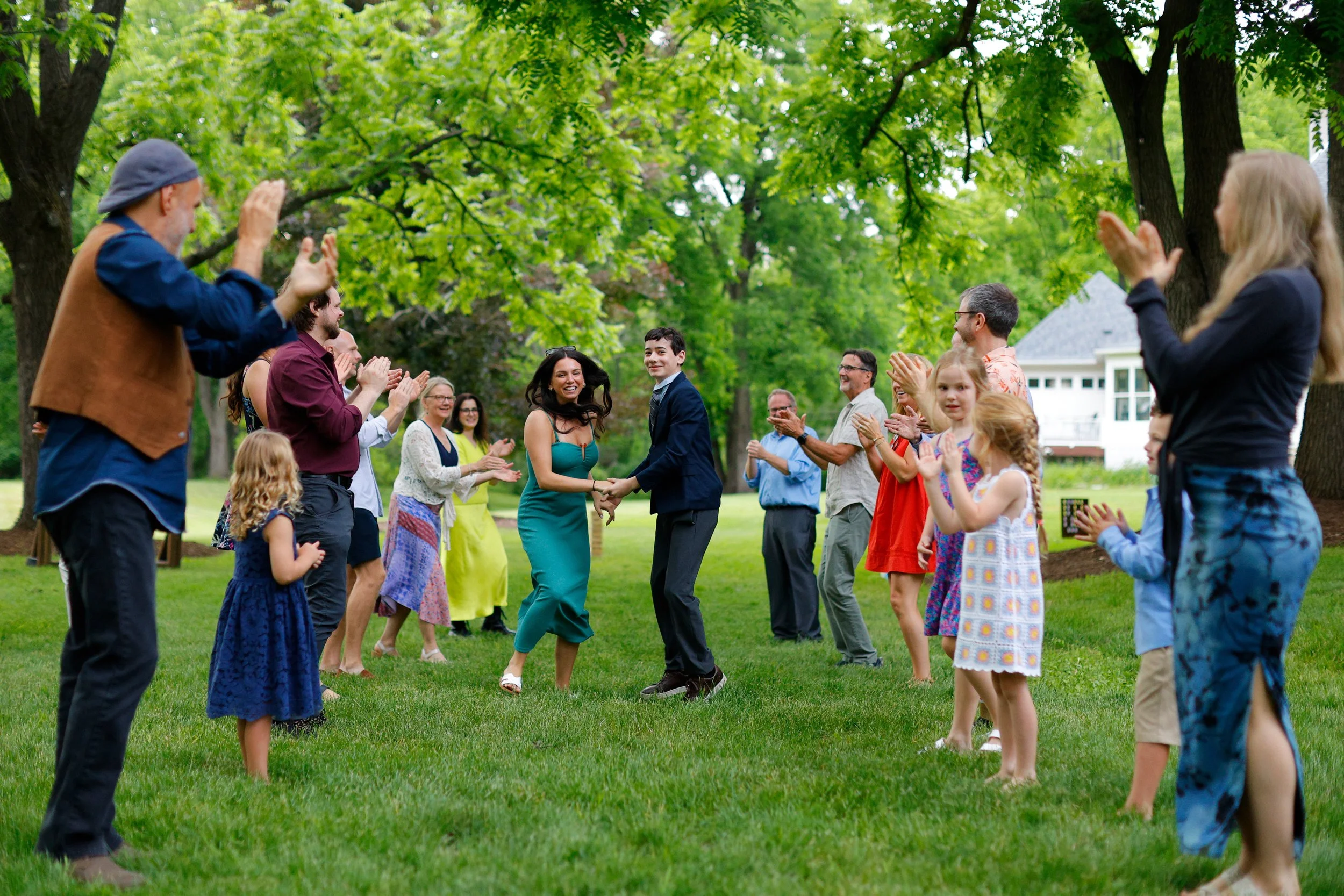 People outdoors on a green lawn under trees, holding hands and clapping, in a celebratory or dance circle, with a smiling couple at the center.