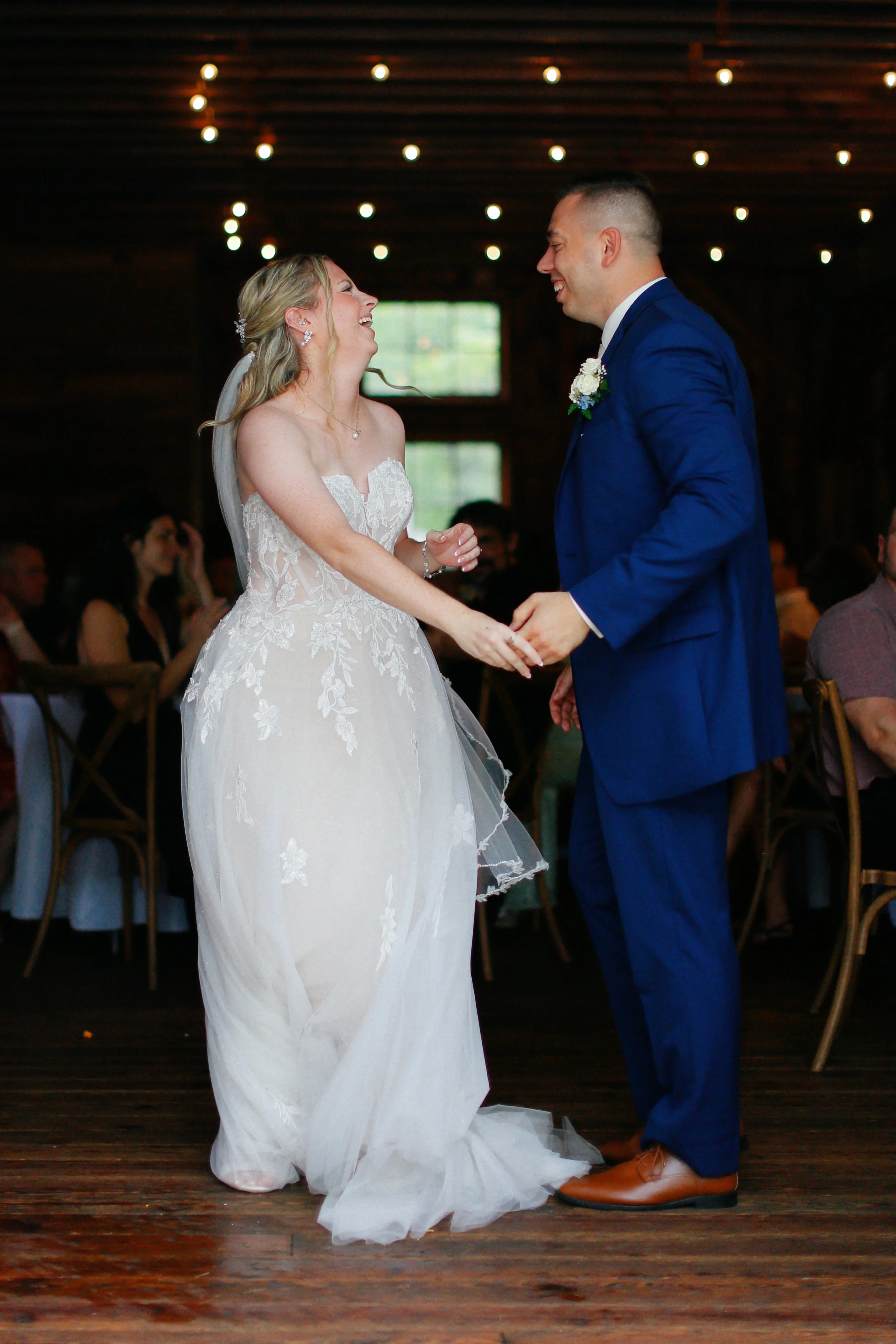 A bride and groom dancing and laughing at their wedding reception in a rustic venue, holding hands and looking joyful.