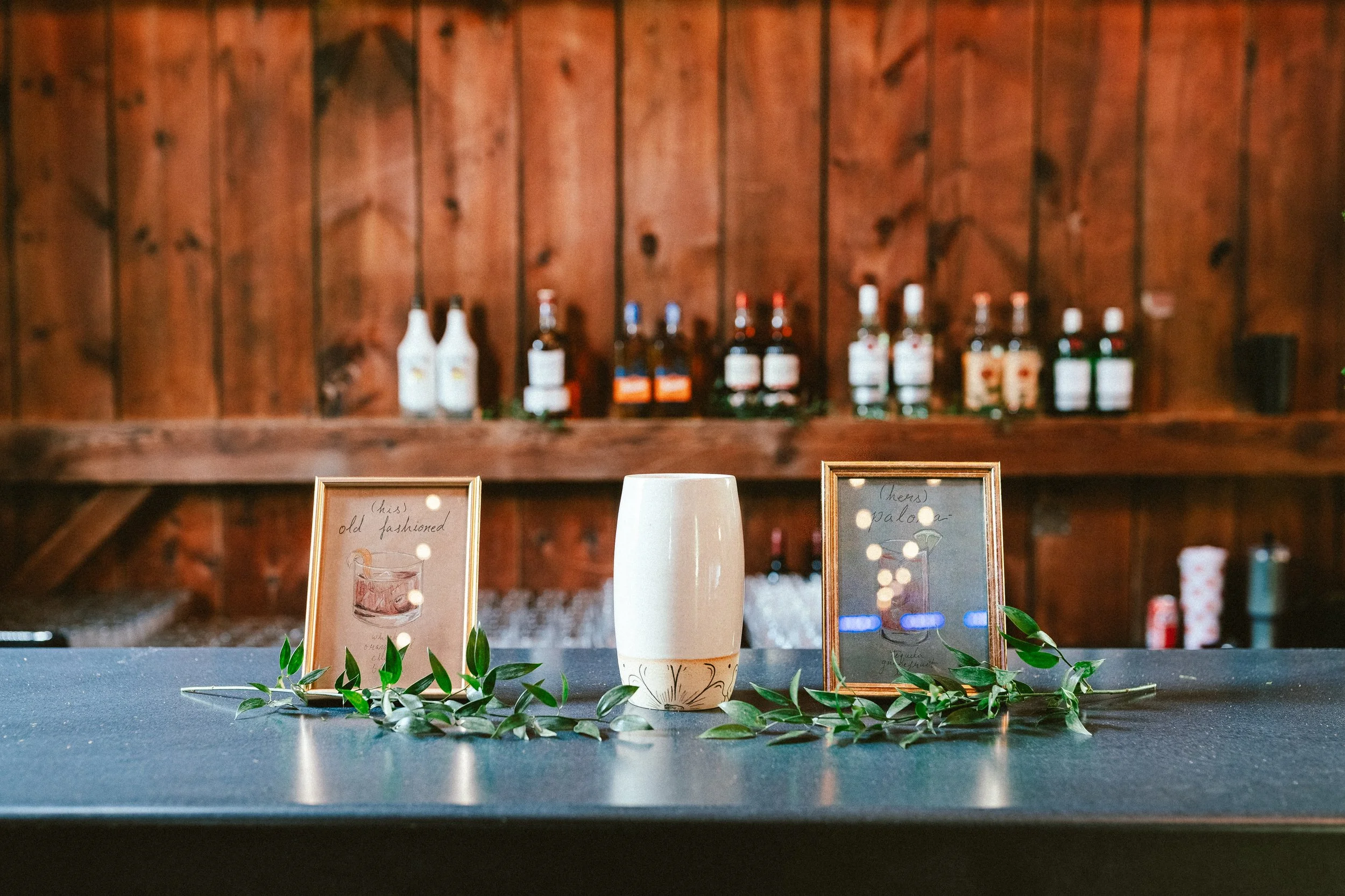 A table with framed drink menu cards, a white ceramic cup, and greenery, with a wooden bar and bottles in the background.