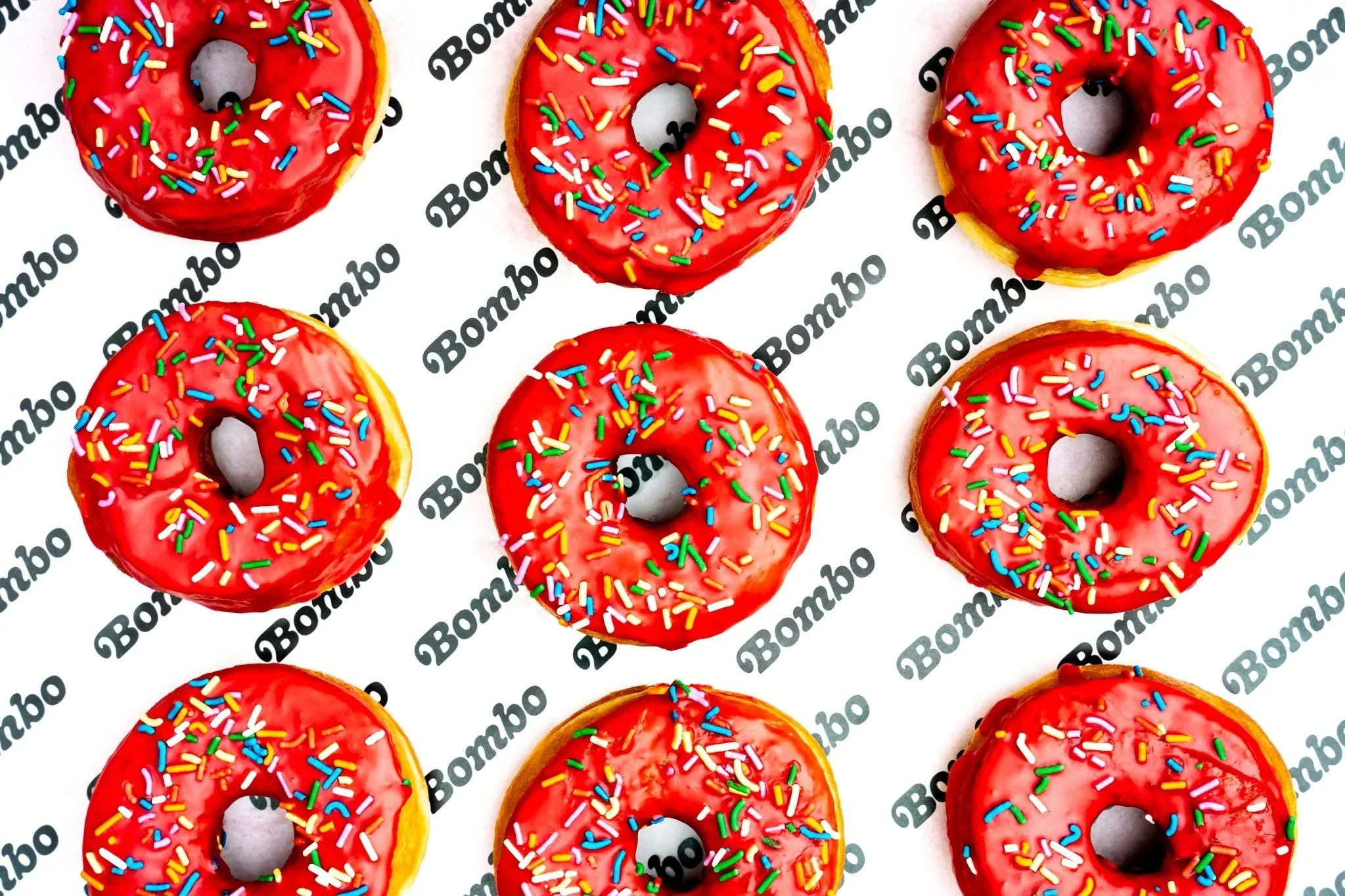 An image of Bombo's famous Wiggum doughnut. A tray of red iced ring doughnuts covered in bright colourful sprinkles, placed on top of black and white Bombo branded paper.