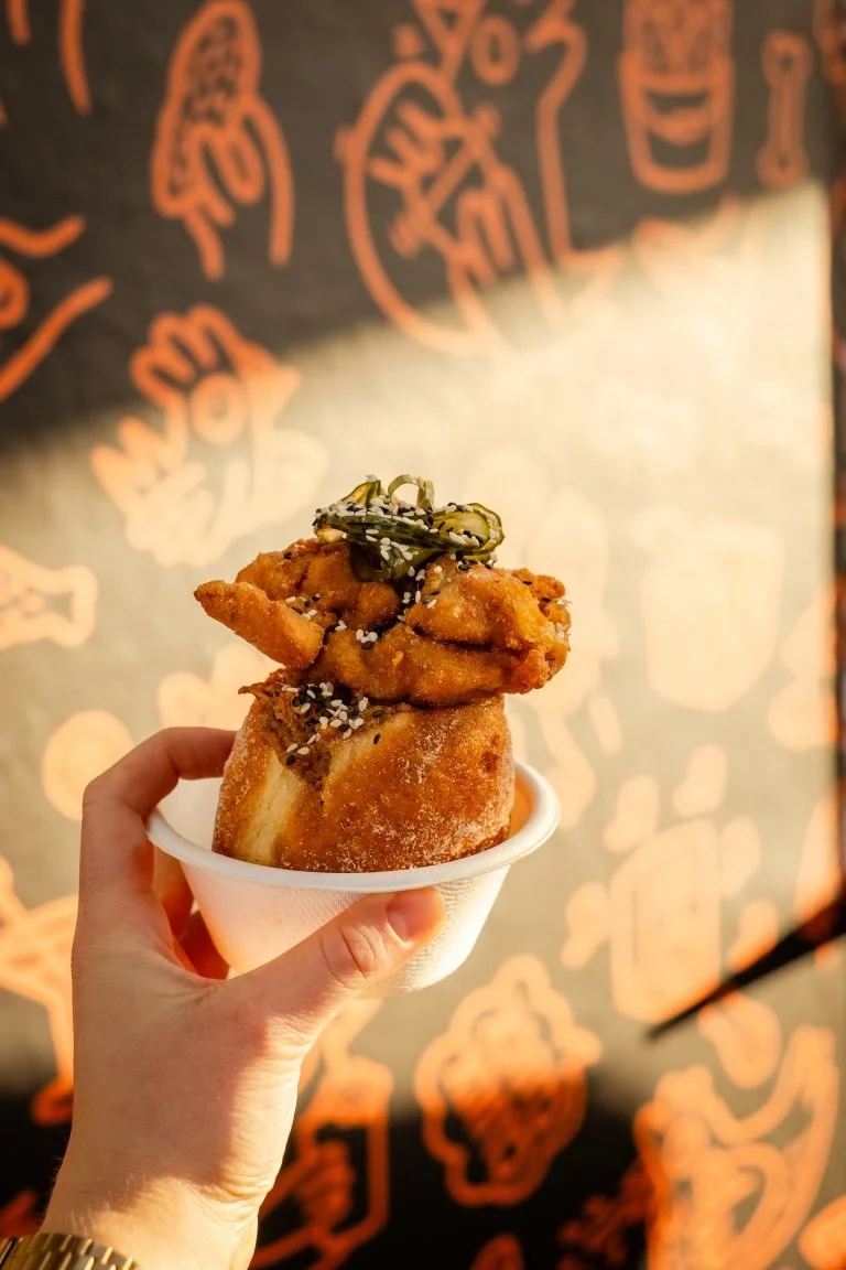 An image of a person's hand, holding a take away bowl, containing a duck filled doughnut topped with a piece of fried chicken and pickles. Yep, that's right, it looks as good as it sounds.