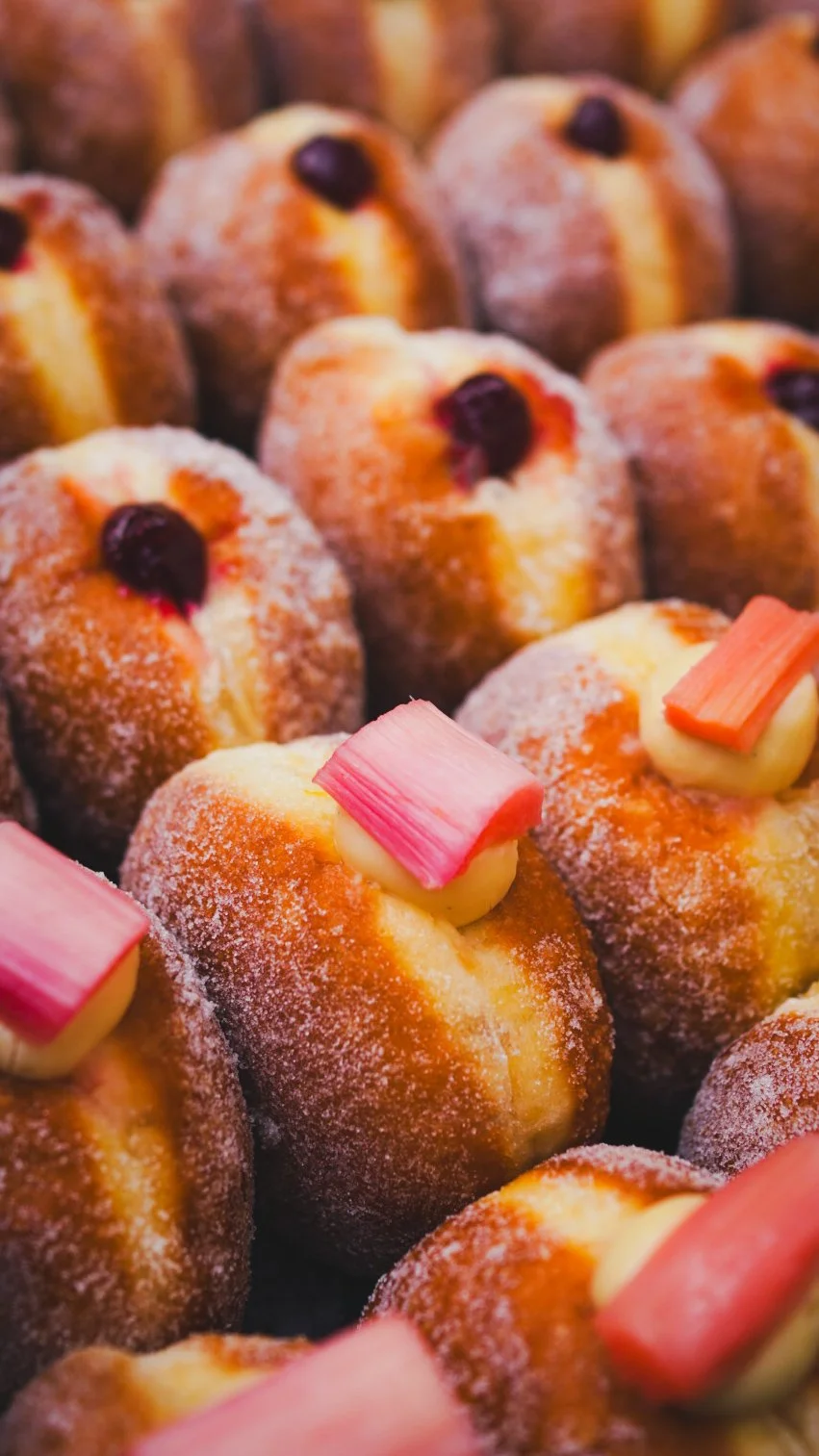 A display of the sexiest doughnuts you've ever seen. Sugar glistens in the light, inviting you to lick the screen. Plump doughnuts sit in neat rows, toped with silky custard and tempting pink slices of rhubarb.
