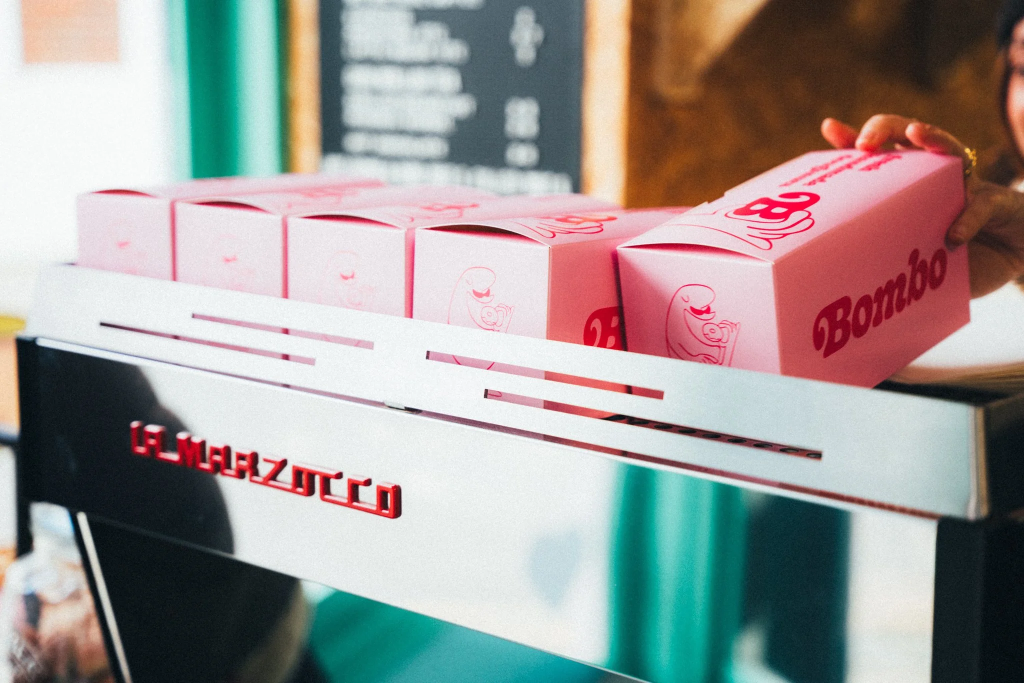 An image of Bombo's pink branded doughnut boxes neatly stacked in one of their retail stores. A hand is reaching in from the right of the image to grab a box. Maybe it's filled with wonderful fluffy doughnutty treats!