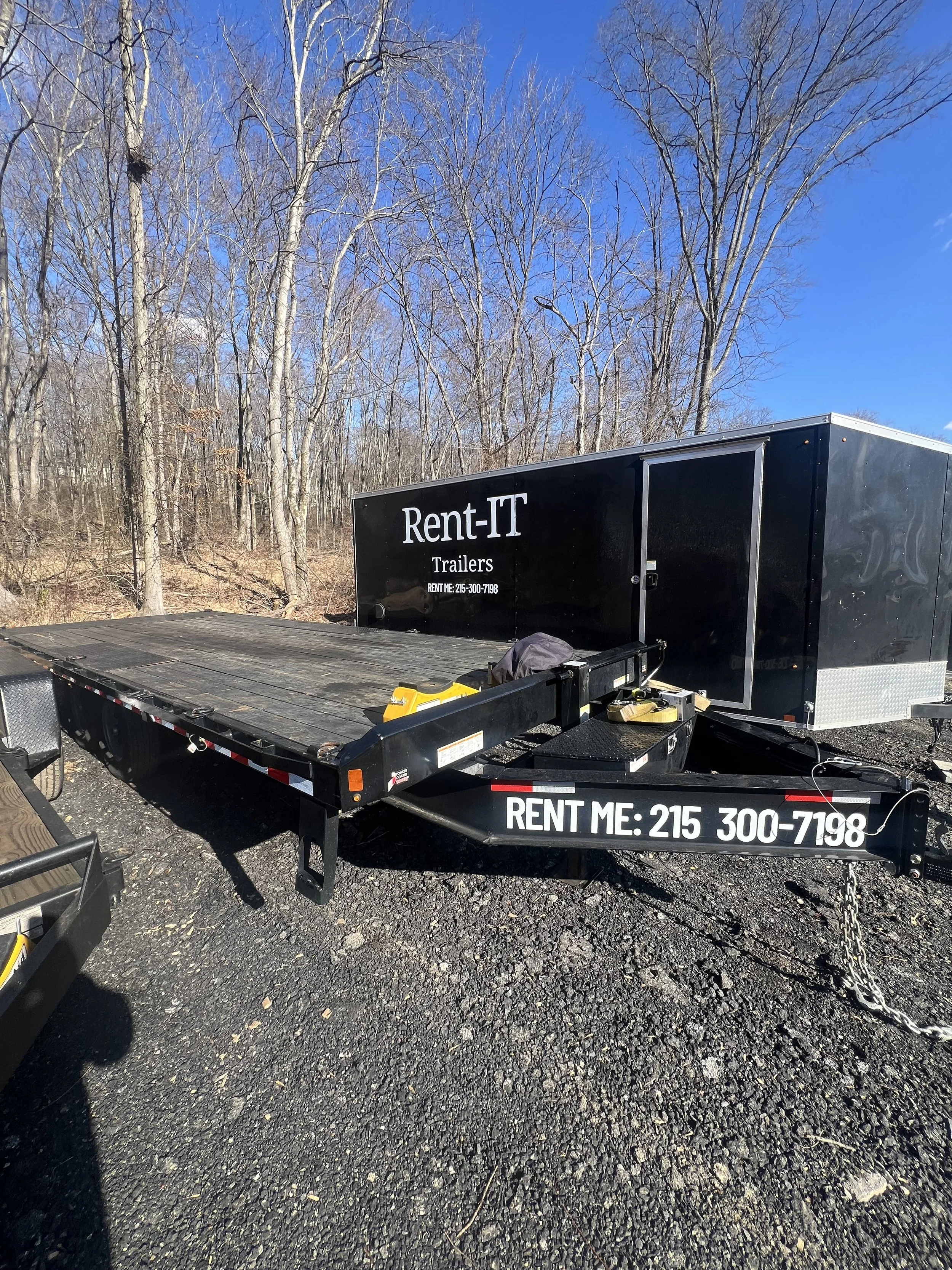 A black flatbed trailer with a black enclosed cargo trailer behind it, both parked on gravel ground next to leafless trees under a clear blue sky. The trailer has white lettering that reads 'Rent-IT Trailers' and a phone number '215-300-7198'.