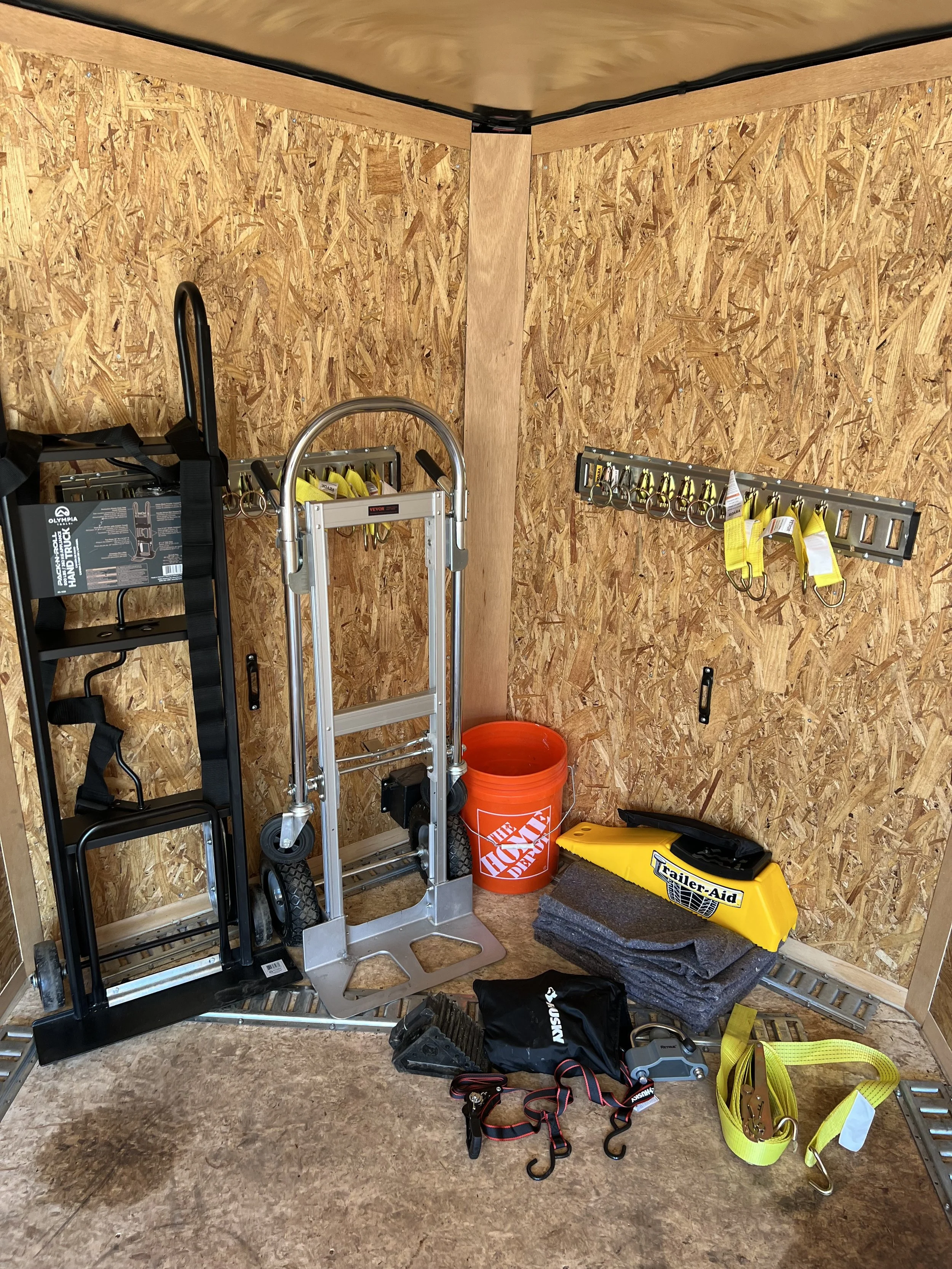 Organized storage corner with tools, straps, and equipment including a yellow trailer aid, orange bucket, and a metal dolly, set against wooden walls.