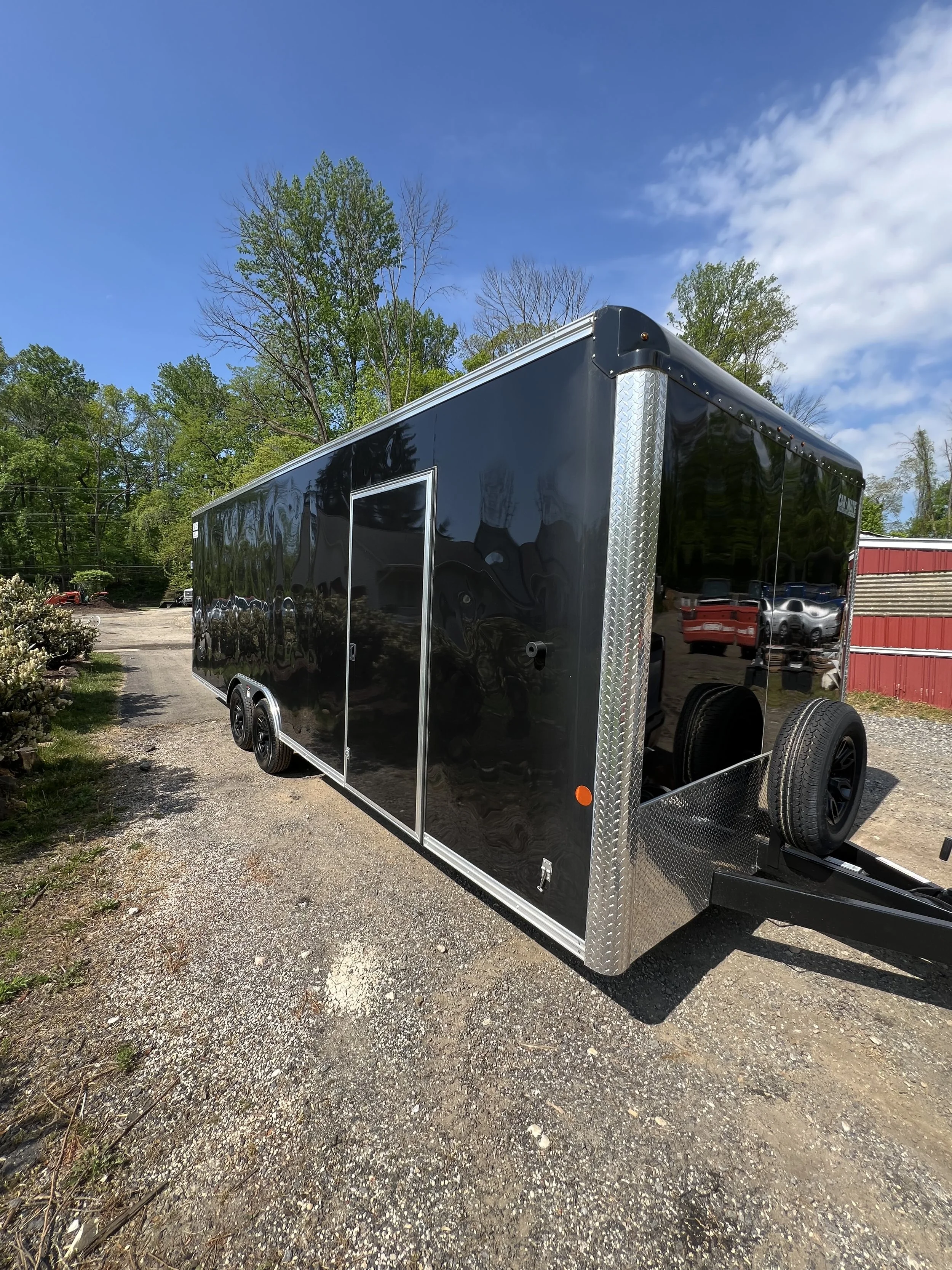 Black enclosed cargo trailer with silver trim parked on gravel ground under a blue sky with trees in the background.