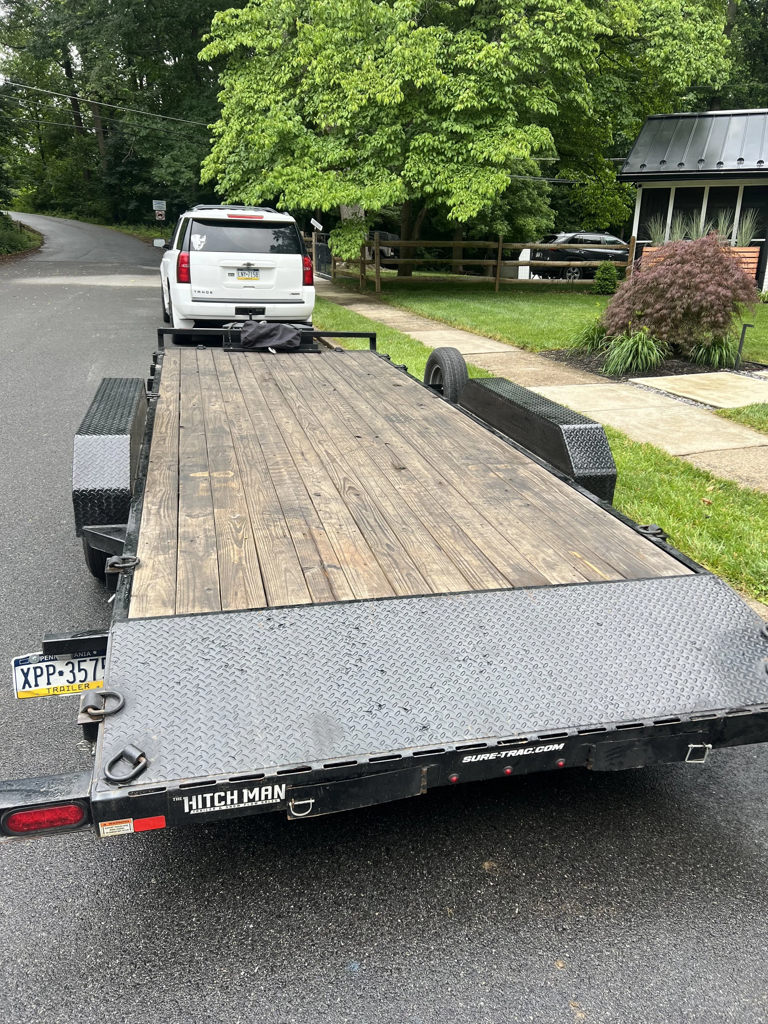 An empty flatbed trailer attached to a vehicle parked on a driveway, with a white SUV parked in front of it. The trailer has a wooden platform and black metal fenders, and is labeled with the brand Hitch Man.