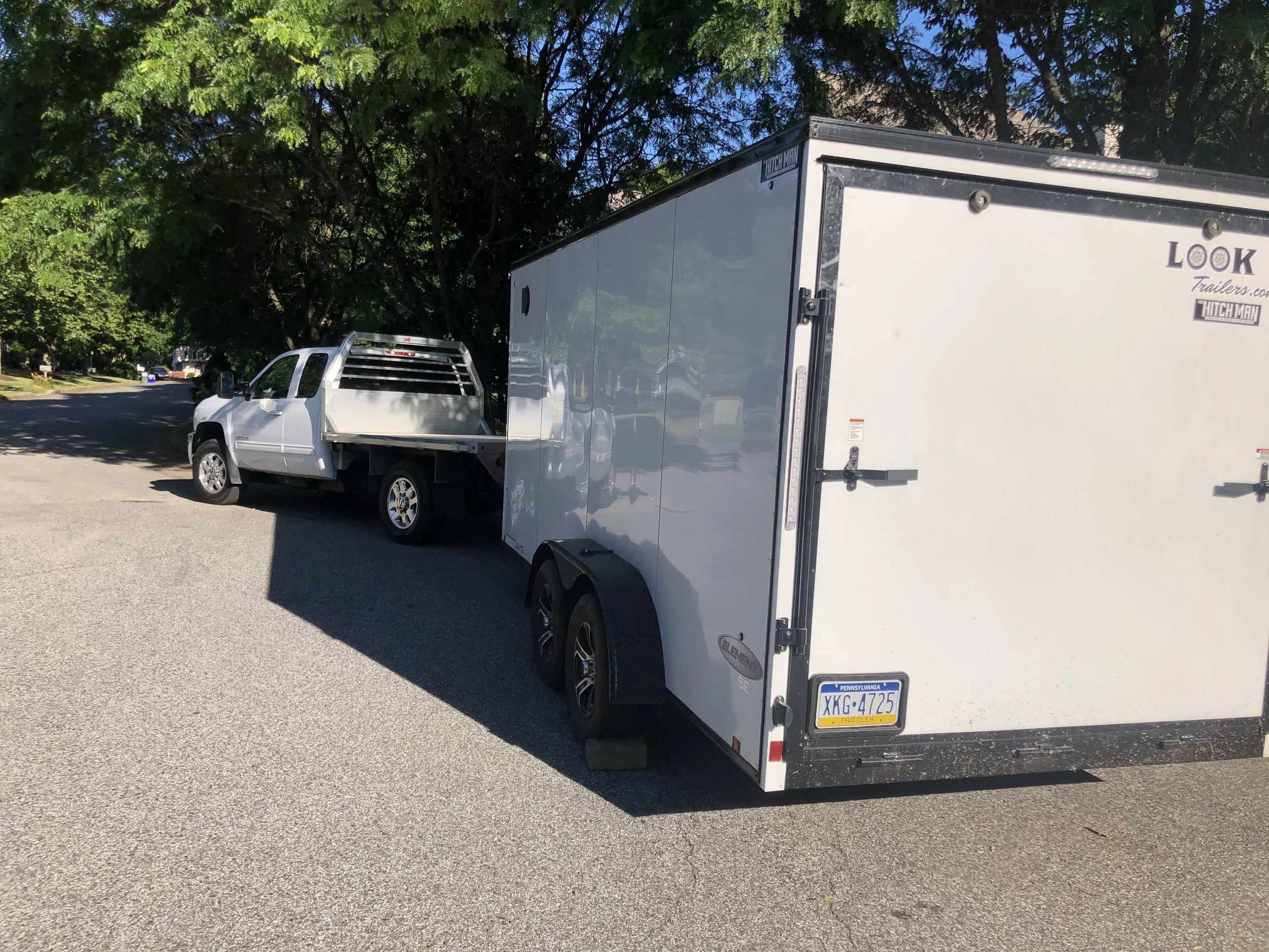 A silver pickup truck towing a white enclosed trailer parked on a residential street with green trees.