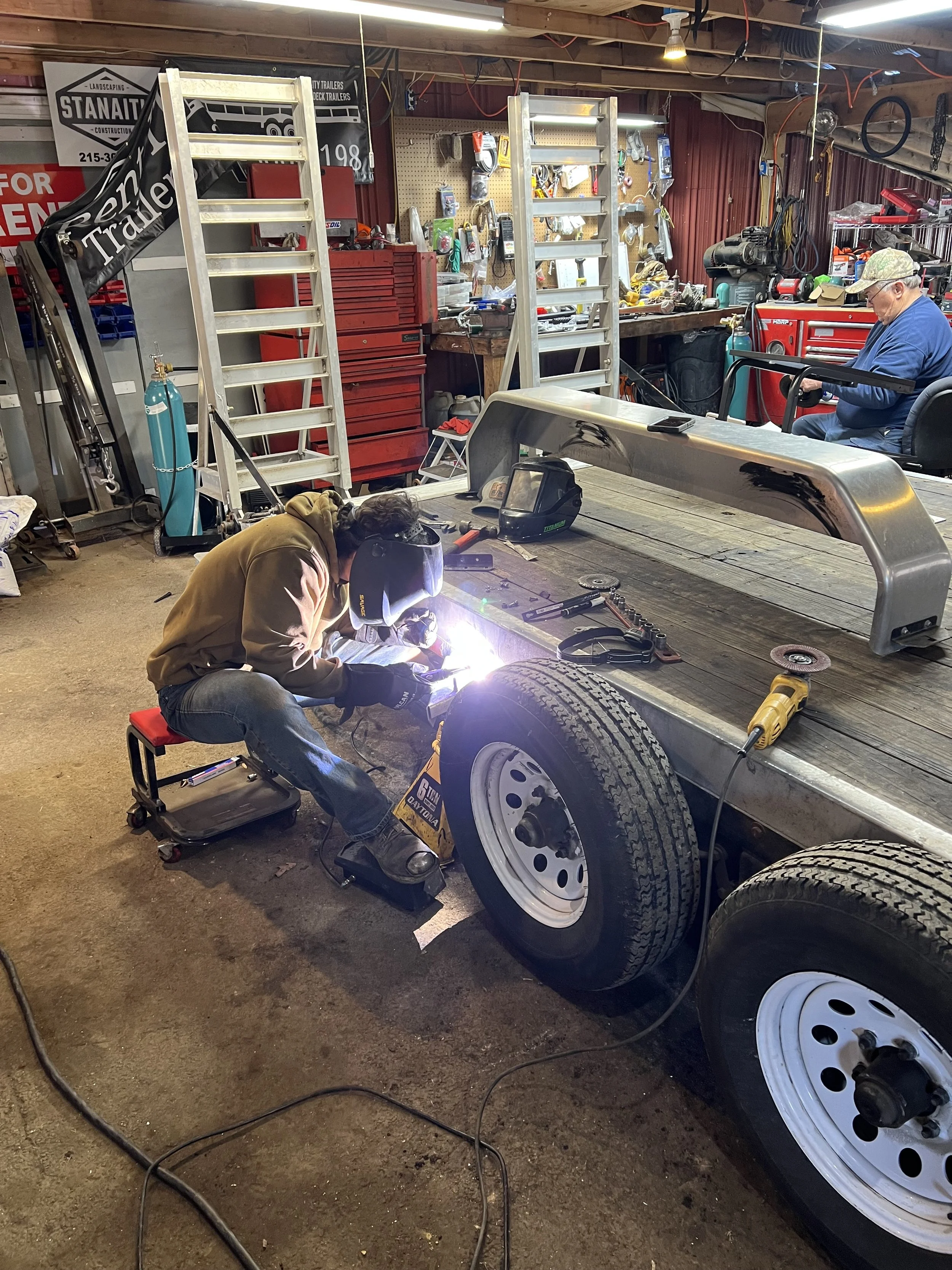 A person welding a wheel on a flatbed trailer in a workshop with tools and ladders.