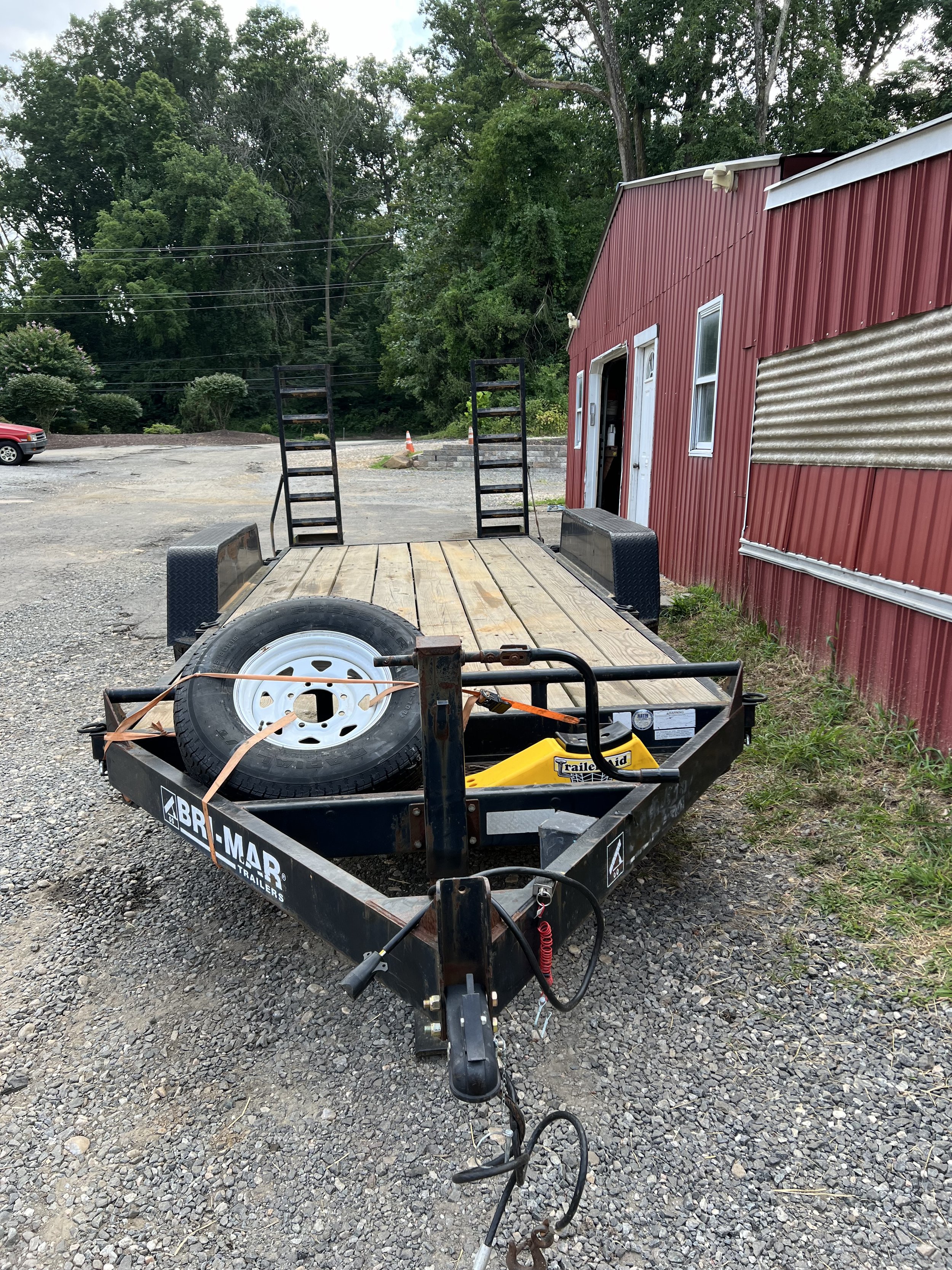 Empty flatbed trailer parked on gravel outside a red building with trees in the background.