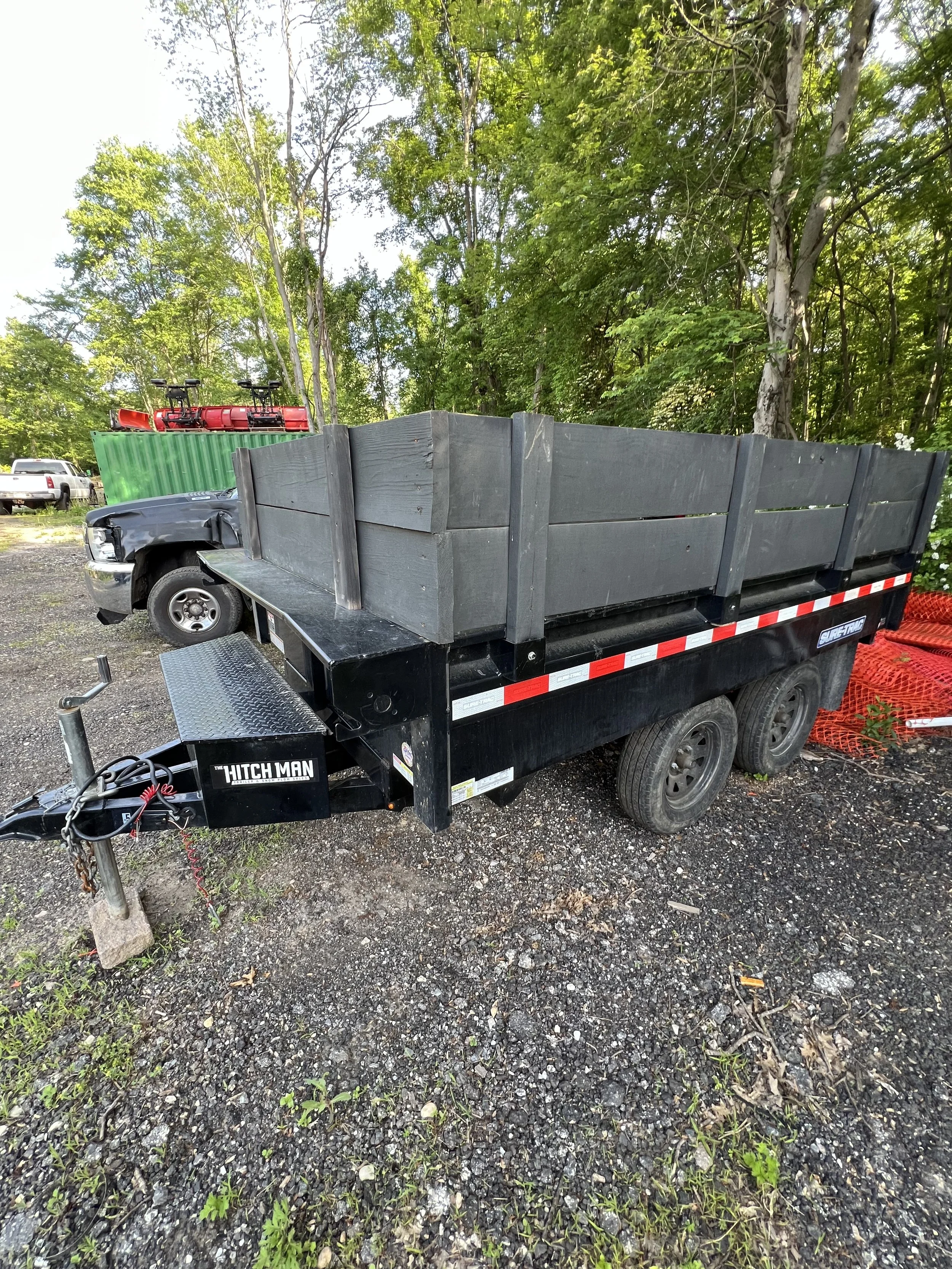 A black utility trailer with a wooden fence, parked on a gravel surface in a wooded area, with a black truck and a green shipping container in the background.