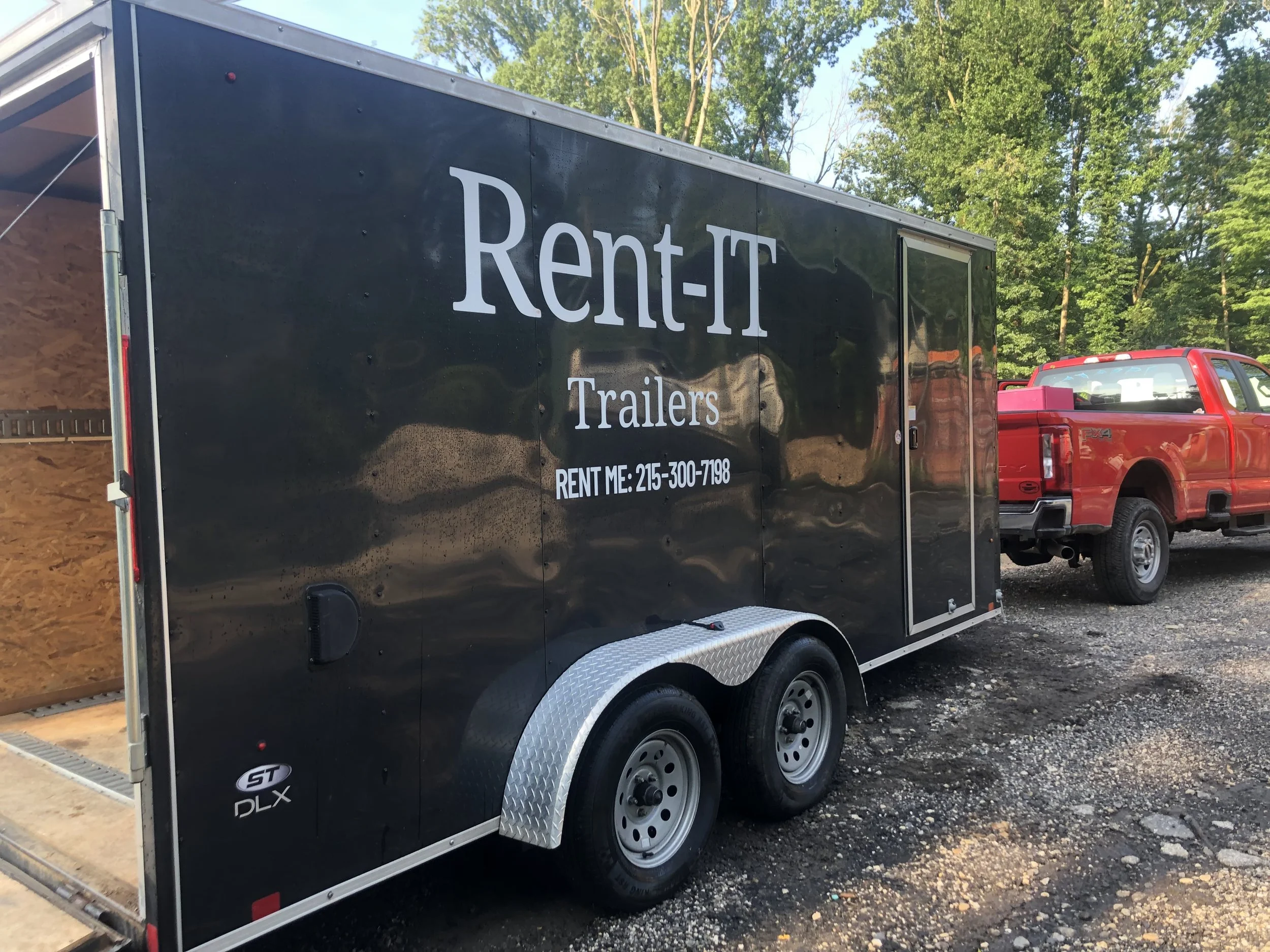 A black trailer with the words 'Rent-IT Trailers' and a phone number on its side, parked on a gravel lot next to a red pickup truck.