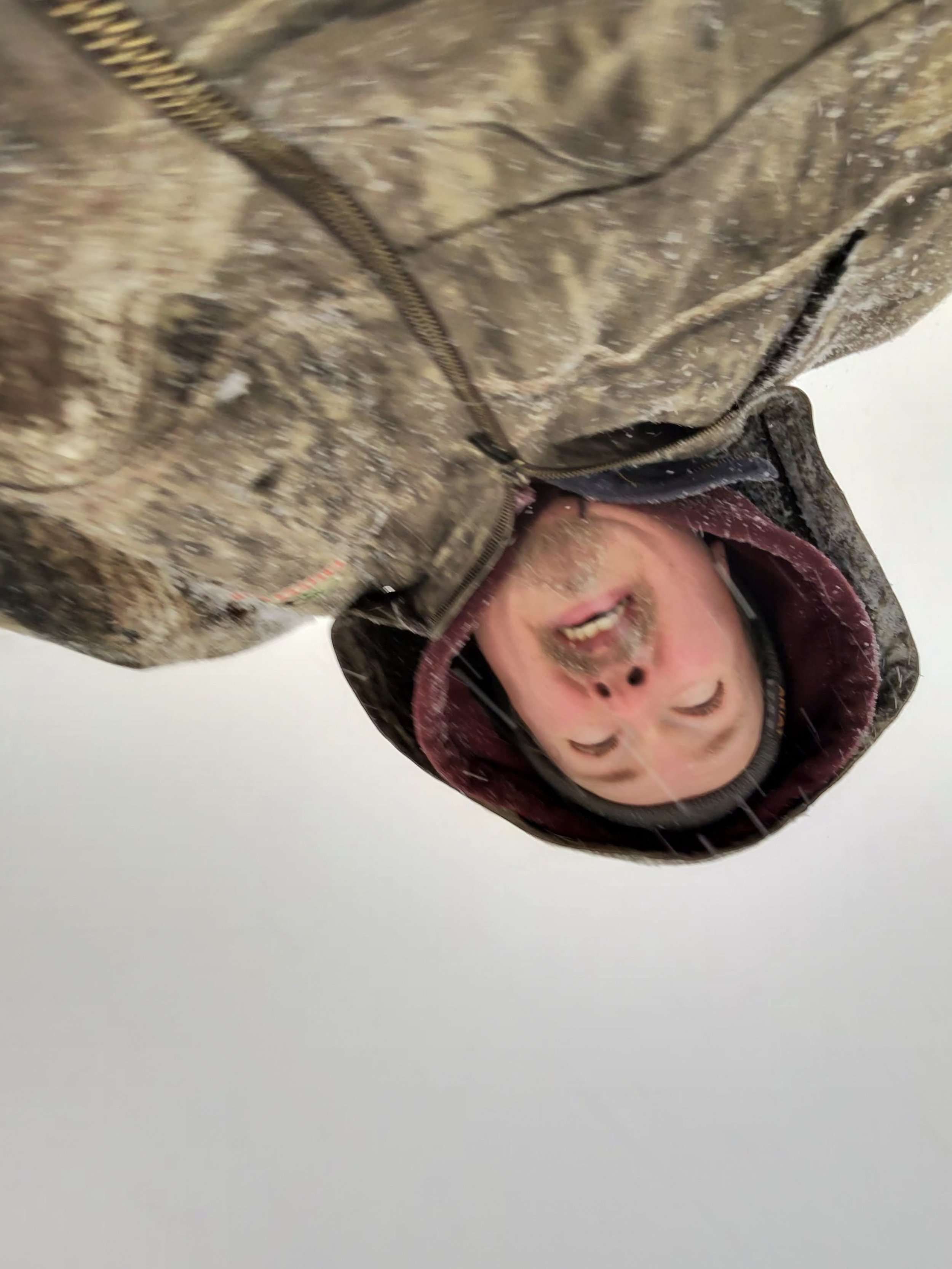 A person wearing a camouflage jacket and a hoodie, smiling with their eyes closed, upside down in a snowy environment.