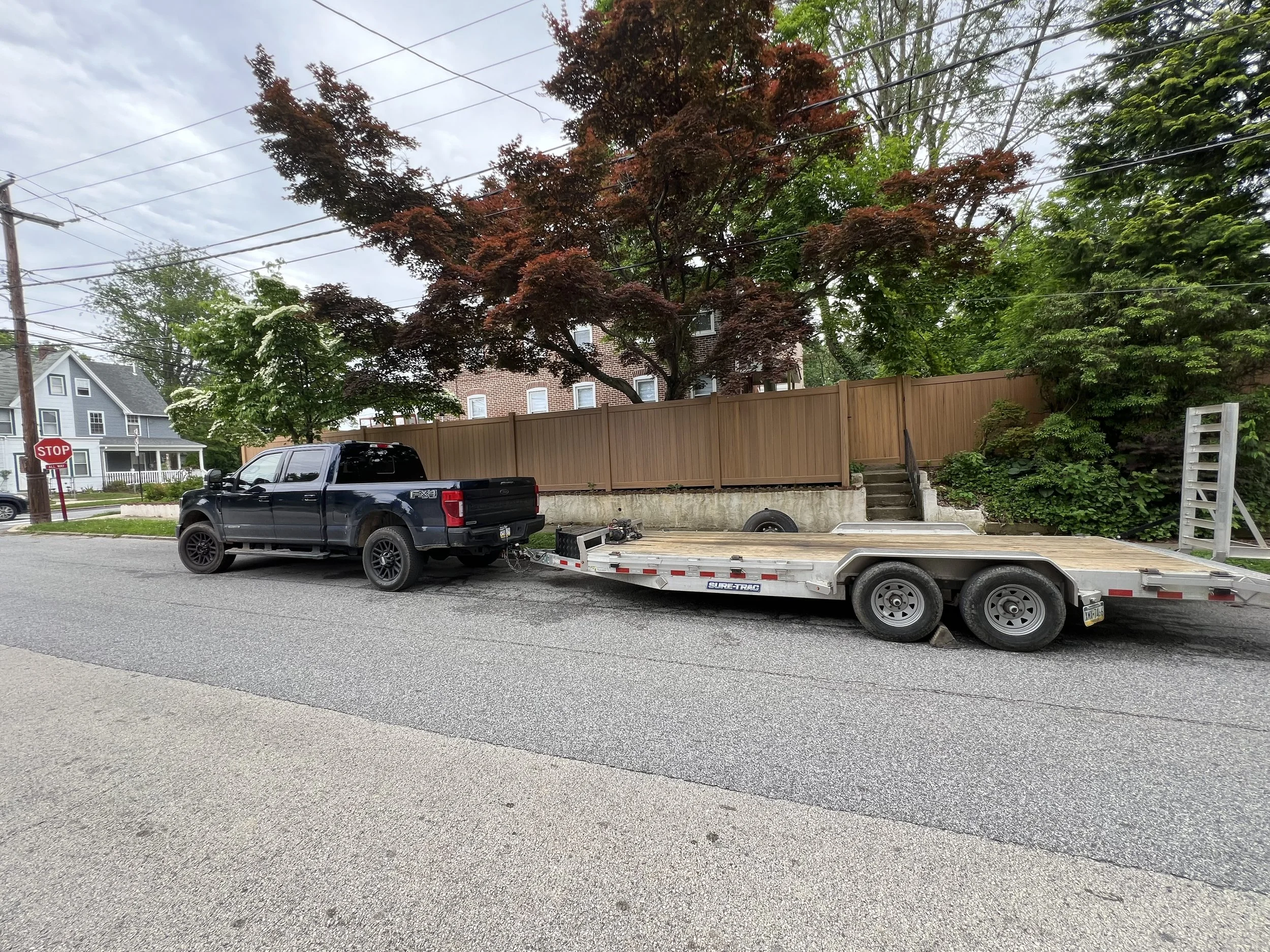 Black pickup truck towing a flatbed trailer parked on a residential street with trees and houses in the background.