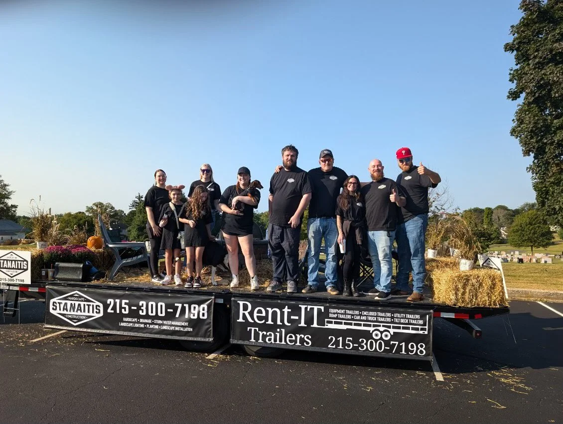 Group of people standing on a float decorated with hay bales and pumpkins, at an outdoor event, with a clear blue sky and trees in the background.
