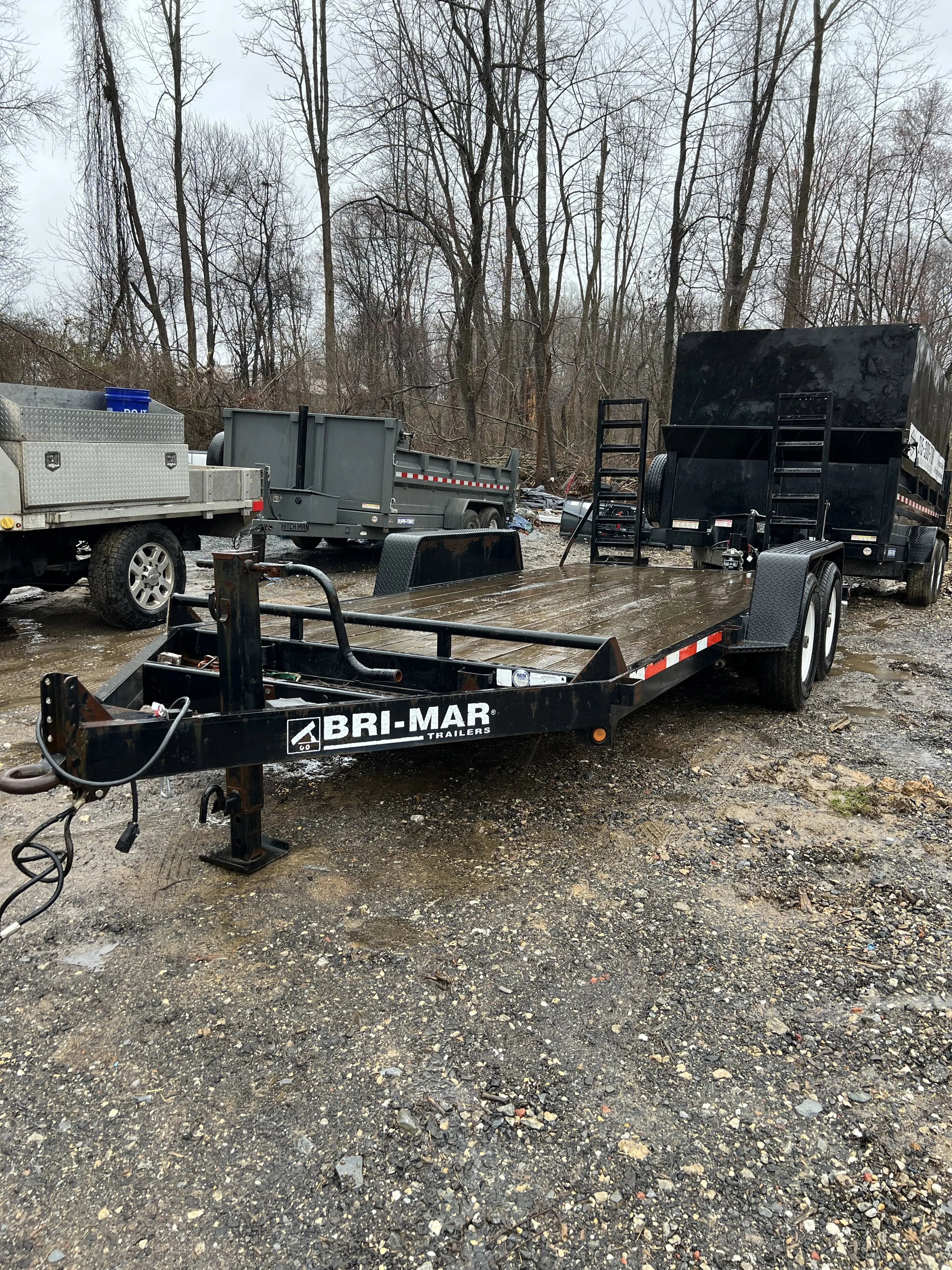 Black flatbed trailer with the label BRI-MAR, parked on a muddy gravel lot outdoors, with other trailers and leafless trees in the background.