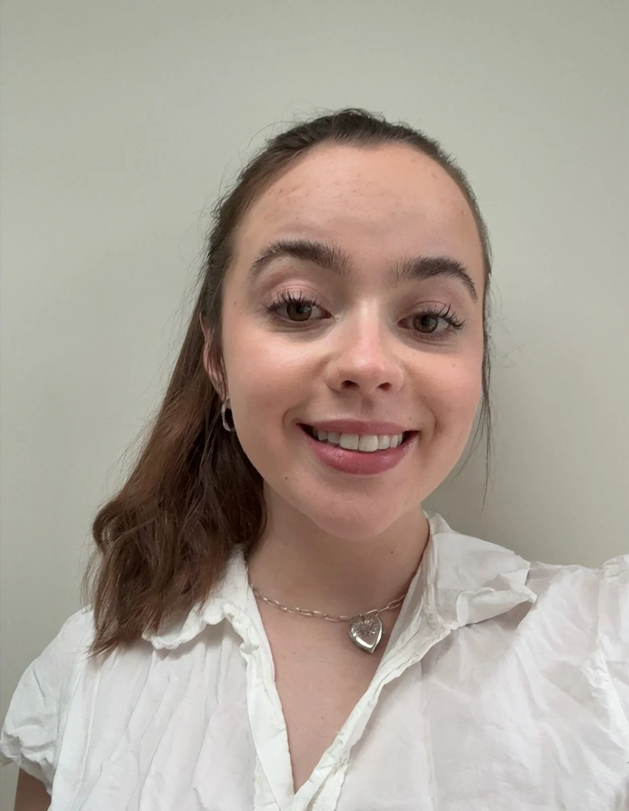 A young woman with brown hair, light skin, and brown eyes smiling, wearing a white blouse and a silver heart necklace, in front of a plain light-colored wall.