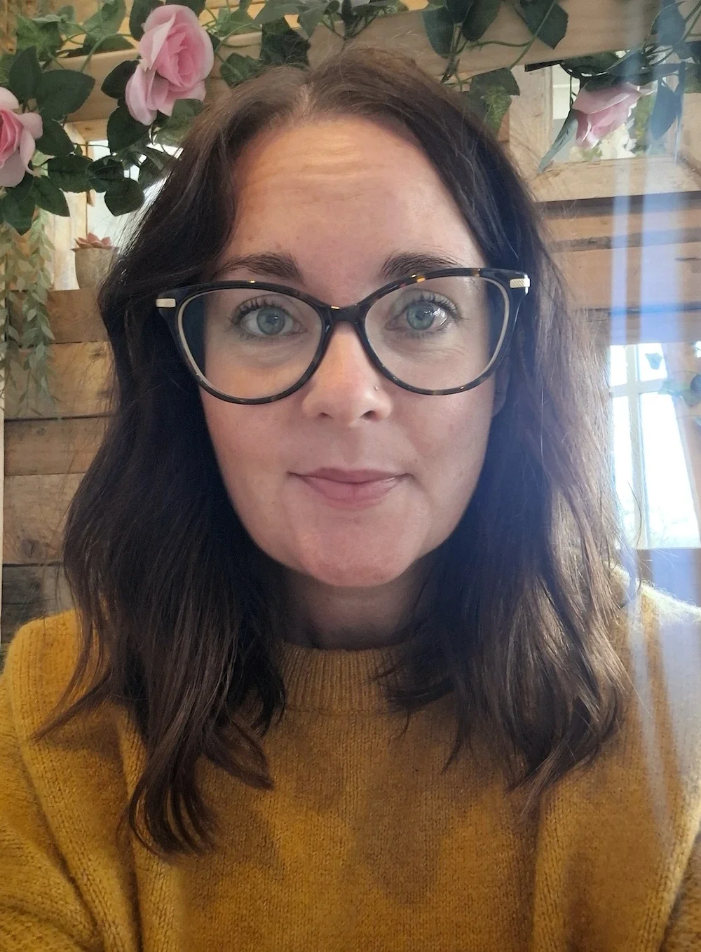A woman with curly dark brown hair, wearing black-rimmed glasses and a mustard yellow sweater, sitting indoors near a wooden wall with pink roses and greenery in the background.
