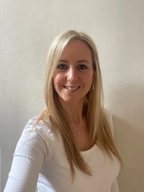 A young woman with long blonde hair smiling in front of a plain light-colored wall.