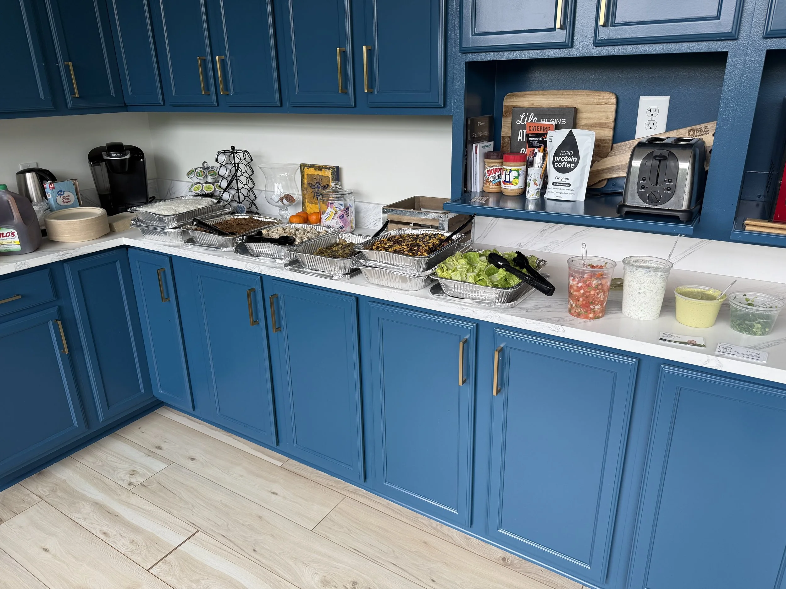A kitchen countertop with blue cabinets and various food items and appliances, including a coffee maker, toaster, and containers with food, along with some books and decorative items.