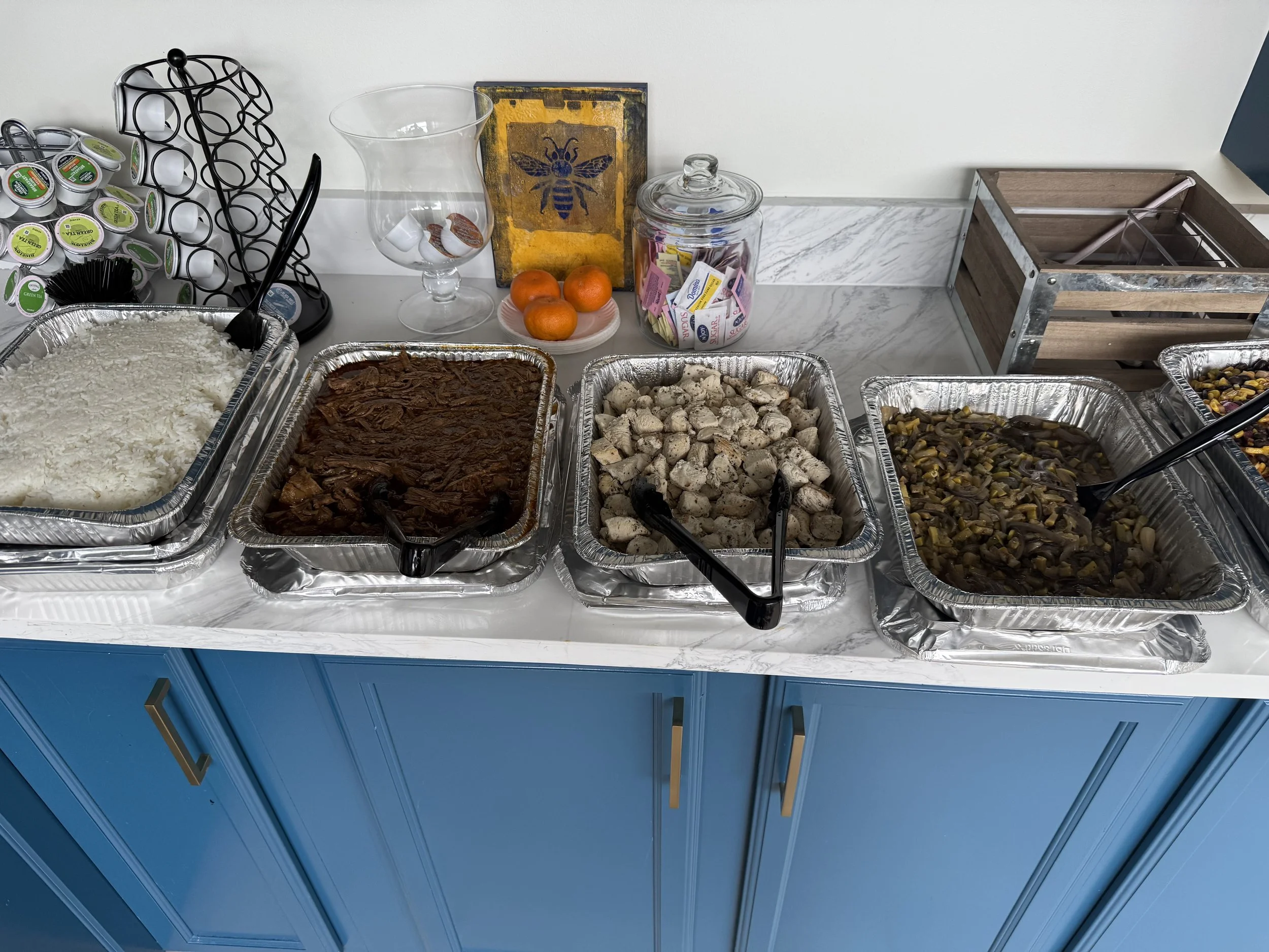 Buffet of various dishes in aluminum trays, including rice, shredded meat, diced chicken, and vegetable medley, placed on a white marble counter with condiments and decorative items behind.