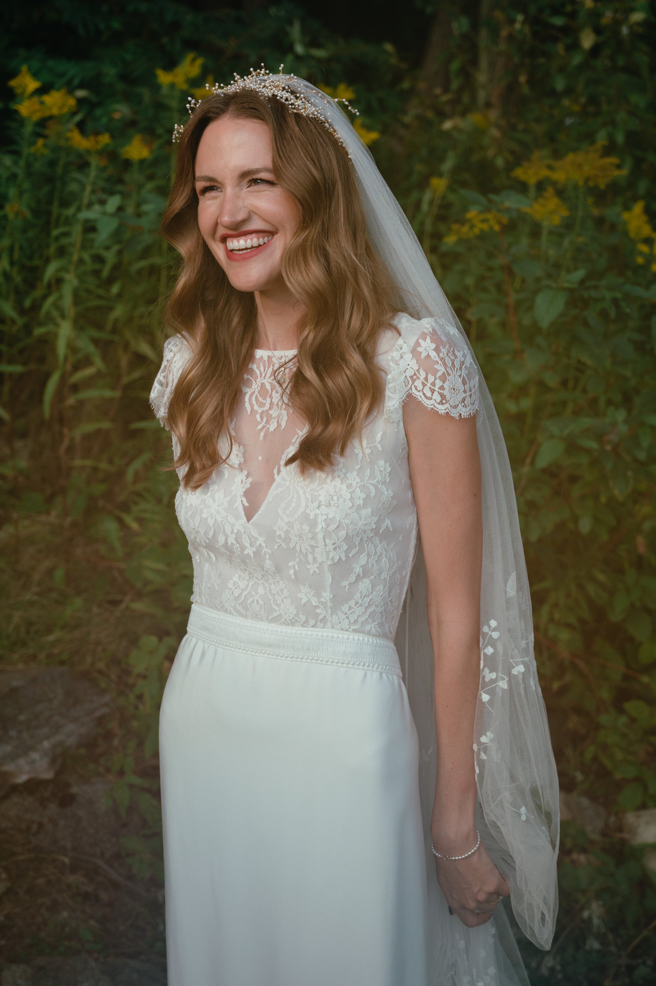 A woman in a wedding dress smiling outdoors with green foliage and yellow flowers in the background.