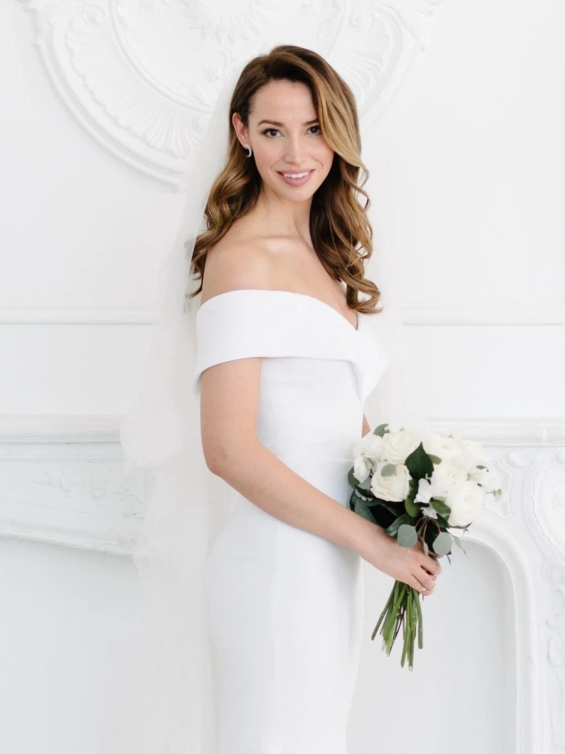 A woman in a white off-shoulder wedding dress holding a bouquet of white roses and greenery, standing indoors against a white ornate wall.