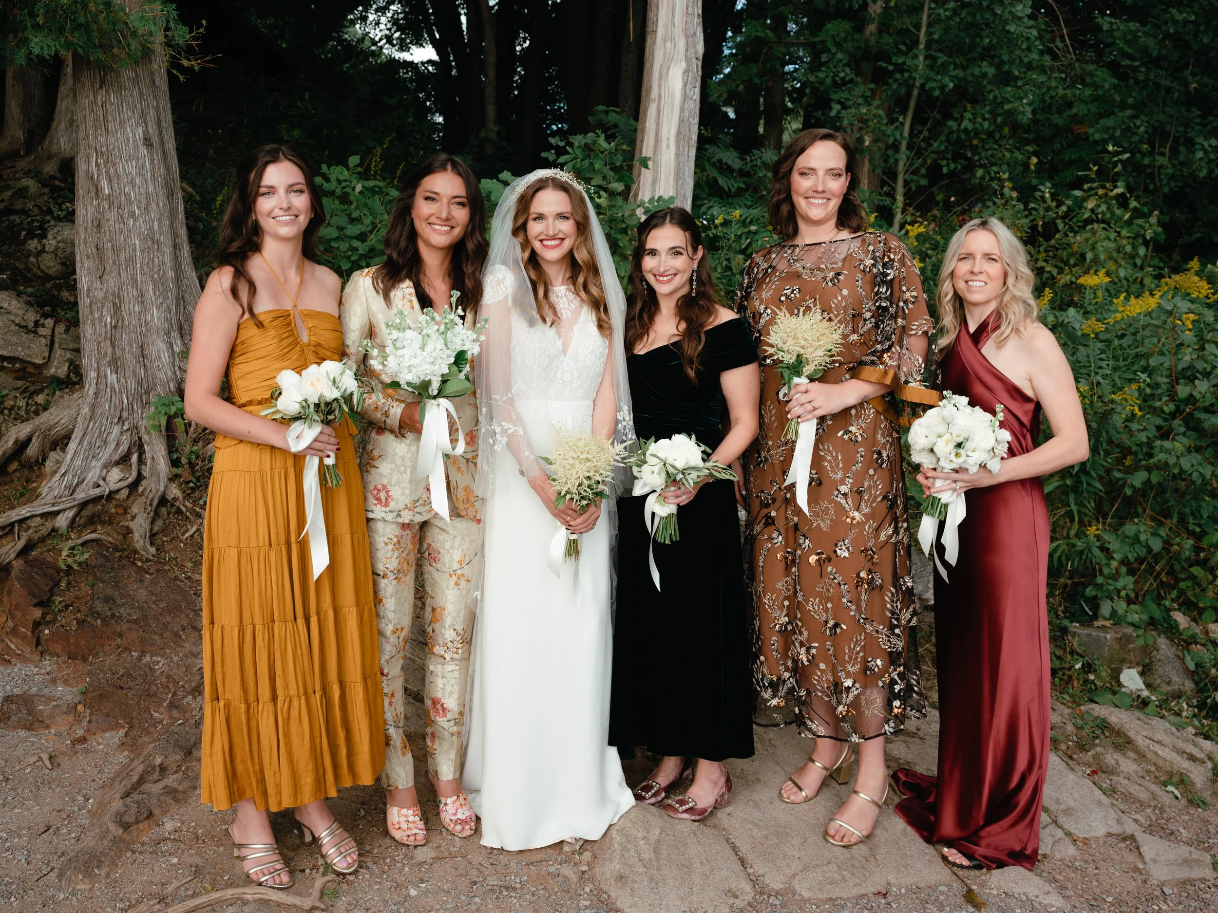 Group of seven women standing outdoors, dressed in colorful dresses, holding bouquets, on a dirt path with trees in the background.