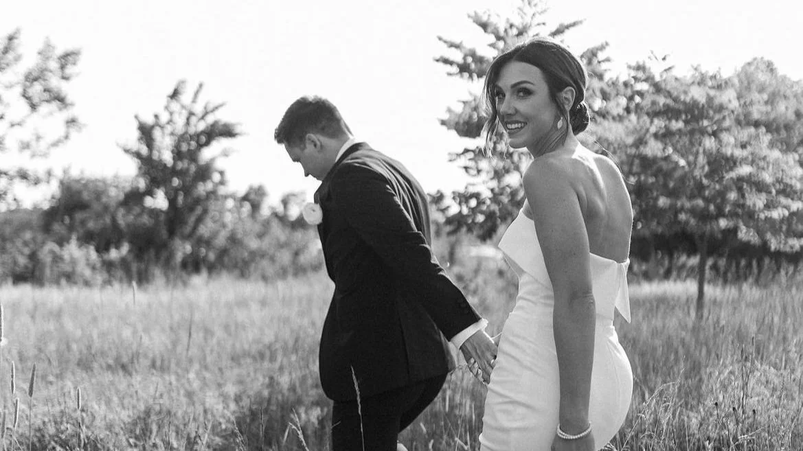 A bride and groom holding hands in a grassy field, with trees in the background, smiling at the camera in black and white.