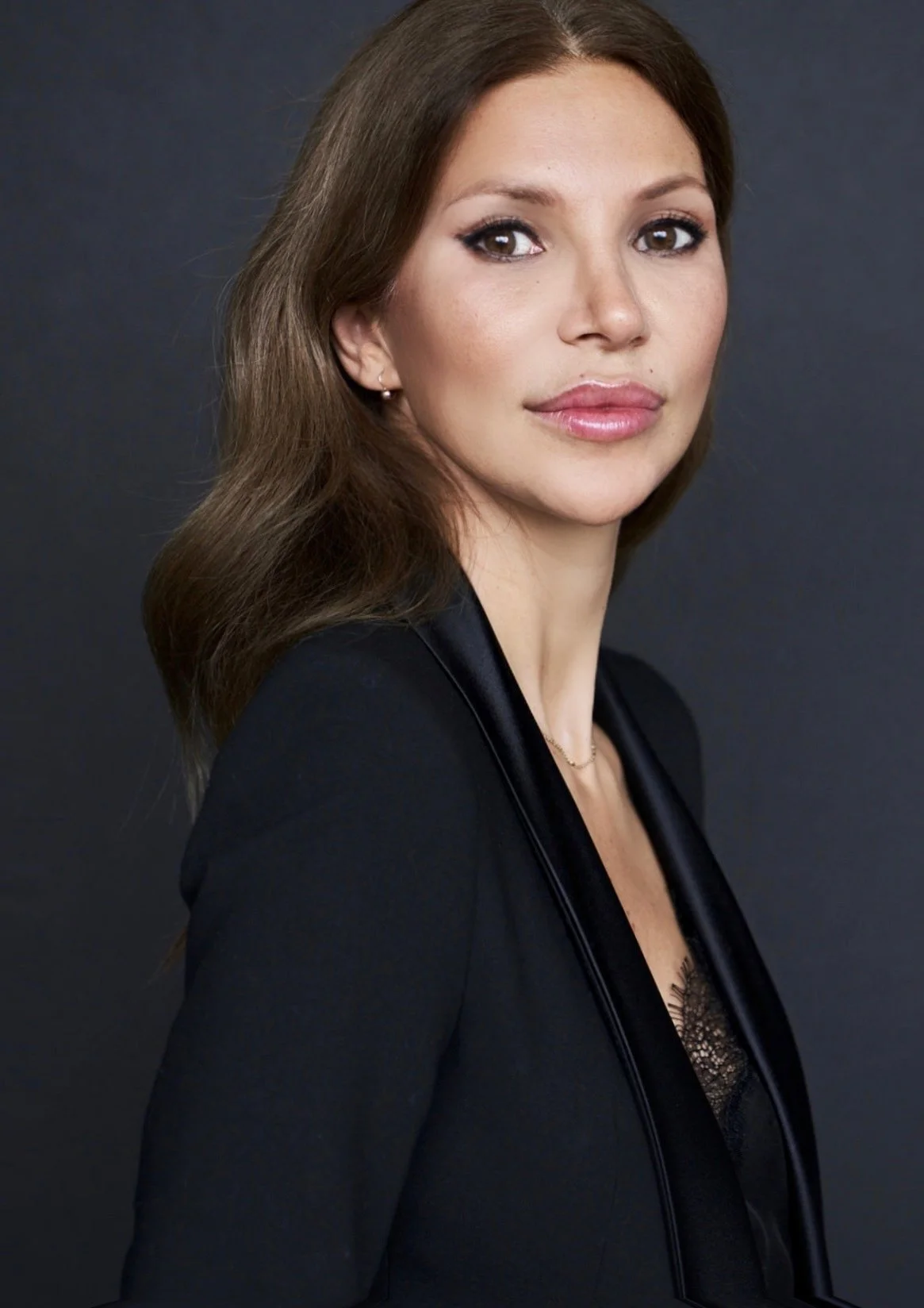 A woman with brown hair, wearing a black blazer with satin lapels and a lace top underneath, posing against a dark background.